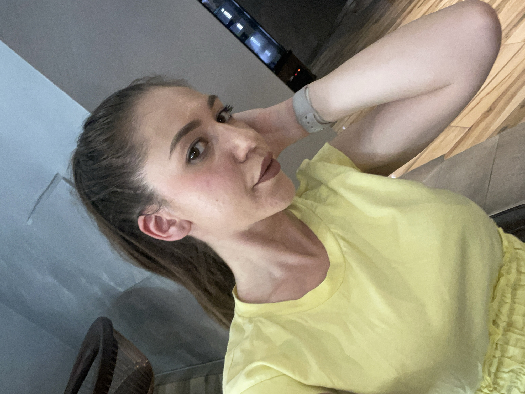 A young woman is captured in a selfie-style photograph, taken indoors in what appears to be a residential setting in Sofia, Bulgaria. She is lying or reclining, with her head slightly tilted and her left arm bent behind her head, resting on it. Her gaze is directed slightly off-camera, and she has a composed expression.
She has light brown hair pulled back into a ponytail, prominent eyebrows, and is wearing makeup including mascara and nude-toned lipstick. Her complexion is fair, with a subtle blush on her cheeks. She is dressed in a plain, light yellow short-sleeved T-shirt. On her left wrist, she wears a light gray smartwatch with a textured band. Her lower body is partially visible, showing bare legs and what appears to be a matching light yellow pleated or ruffled garment, possibly shorts or a skirt, suggesting a coordinated outfit.
The background features a light wooden floor and dark gray walls. In the upper background, there is a dark, sleek electronic device, possibly a television or soundbar, with small illuminated indicators. Another dark piece of furniture, possibly a chair or table, is visible in the lower left. The lighting is soft and ambient, suggesting an indoor evening setting. No visible text is present in the image.