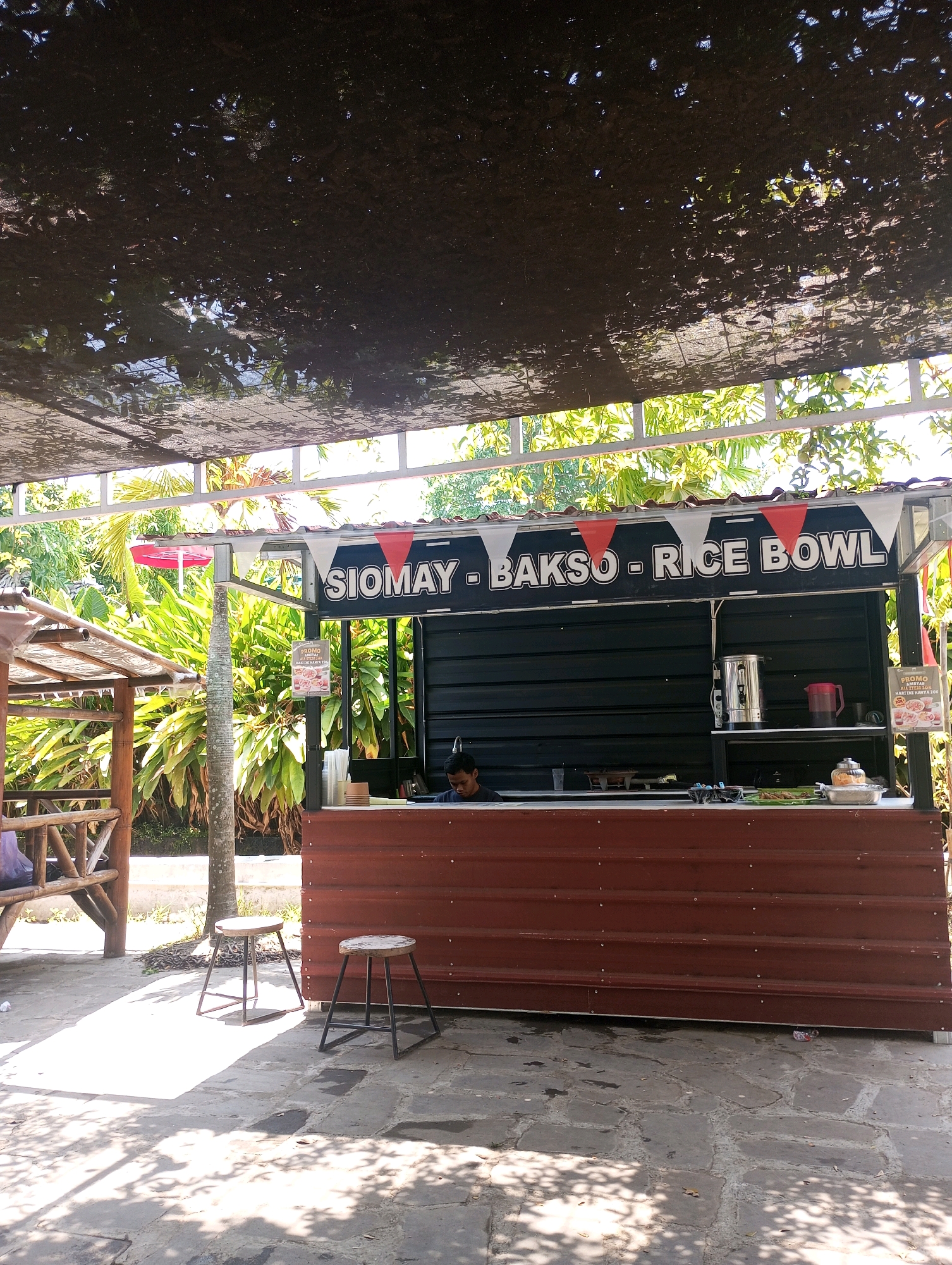 The image depicts an outdoor food stall with a dark corrugated metal back and a brown ribbed front. Above the counter, a banner reads "SIOMAY - BAKSO - RICE BOWL". A man, likely an employee, is visible behind the counter. On the counter, there are various items including a silver kettle, a pink pitcher, cups, and food containers. Two simple metal stools with wooden seats are placed in front of the stall on a stone-paved ground, which is partially covered in sunlight and shadow. To the left of the stall, a wooden gazebo-like structure is visible, with bamboo accents. Lush green tropical foliage forms the backdrop. The scene appears to be set in Ponggok, Indonesia, given the context provided, likely a tourist or recreational area given the outdoor setting and the type of food stall.