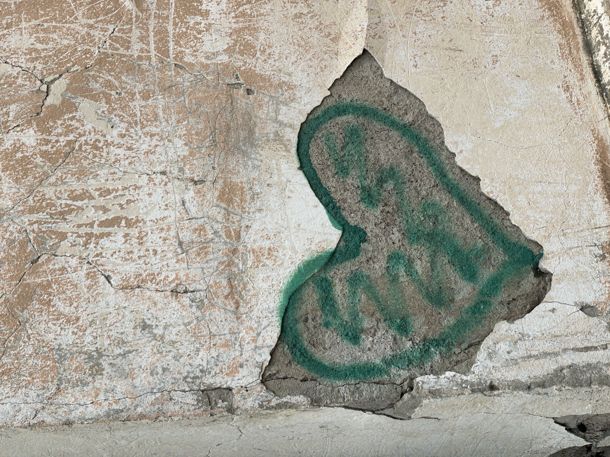 An aged, weathered wall shows significant peeling and decay. A portion of the outer layer has broken away, revealing an underlying surface marked by a circular pattern in faded green spray paint. The wall appears to be concrete or plaster, exhibiting cracks and crumbling textures throughout. The color palette consists predominantly of off-white, beige, and gray tones. The location in Zheleznogorsk, Russia, is inferred but not directly depicted in the image; the image primarily focuses on the textures and damage of the wall itself.