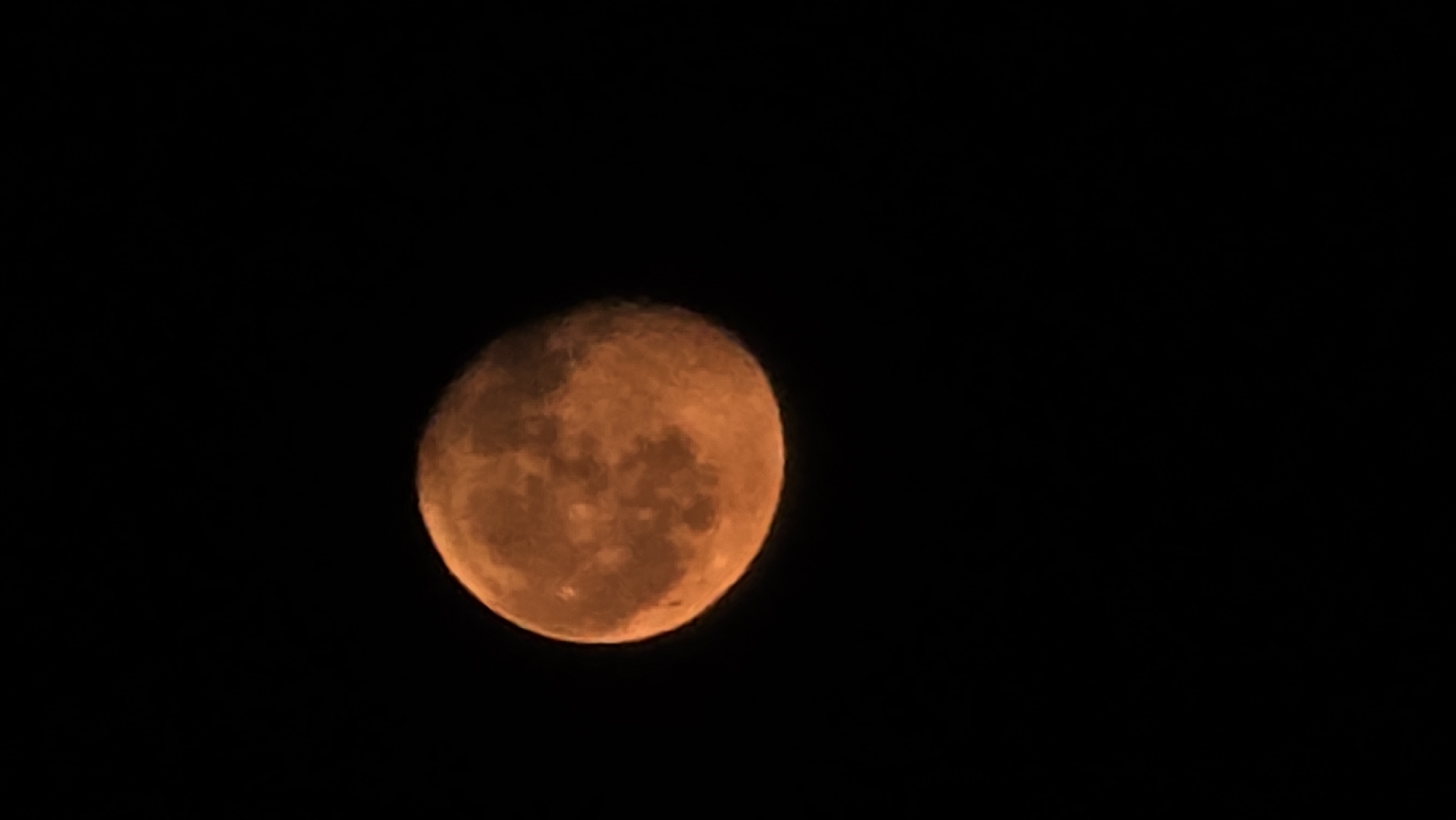 A close-up, low-angle shot captures an illuminated full moon against a black night sky. The moon is a warm, orange hue, with visible surface details suggesting craters and mares. The lower portion of the moon is slightly cropped out of the frame. The image is taken in Krasnodar, Russia.