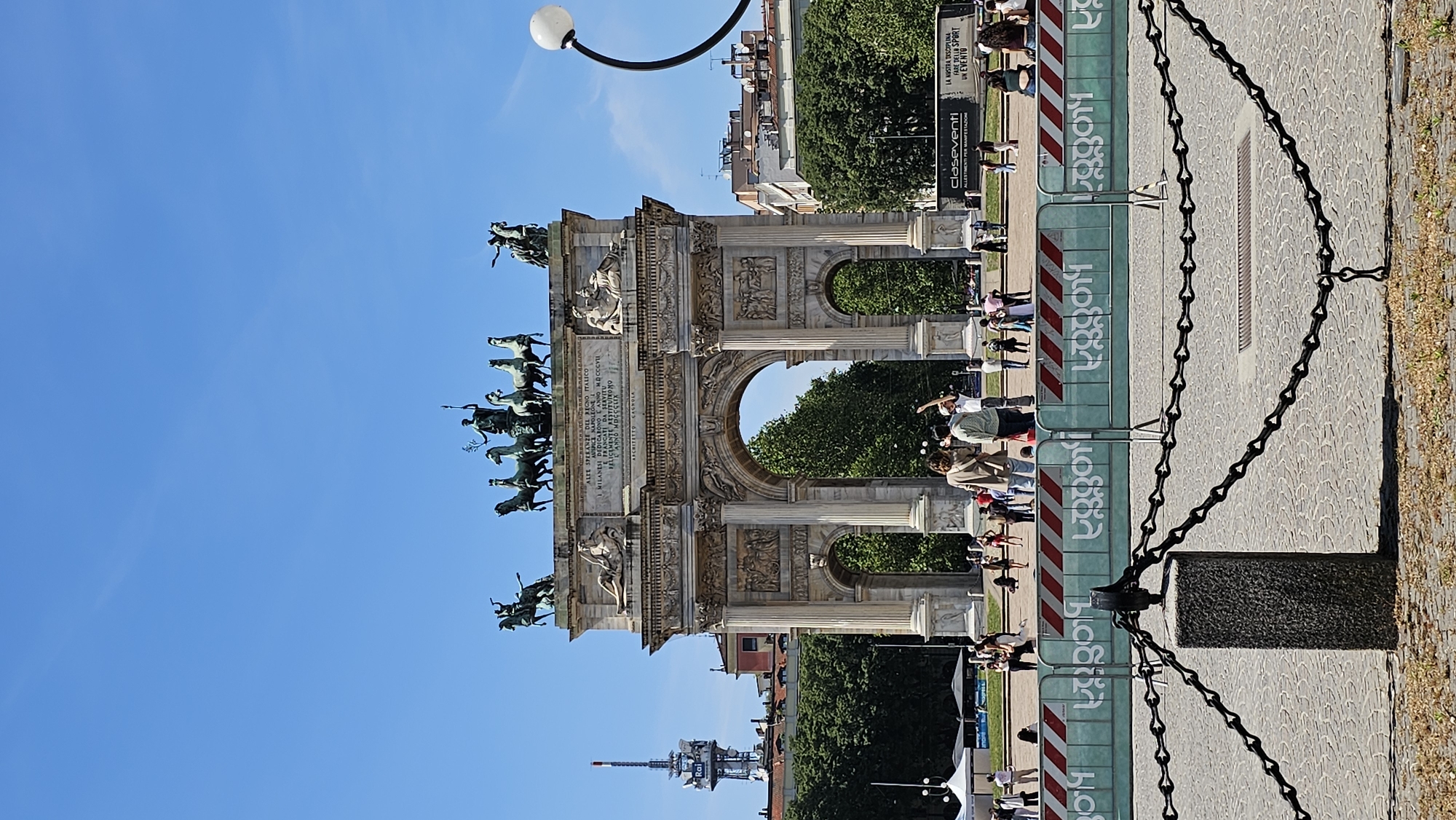 The image shows the Arco della Pace (Arch of Peace) in Milan, Italy, a neoclassical triumphal arch. The arch is made of stone and features sculptures of horses and chariots on top. In front of the arch is a pedestrian area with people walking around. A chain-link fence is in the foreground. Buildings and trees are visible in the background, along with a telecommunications tower. The sky is blue.