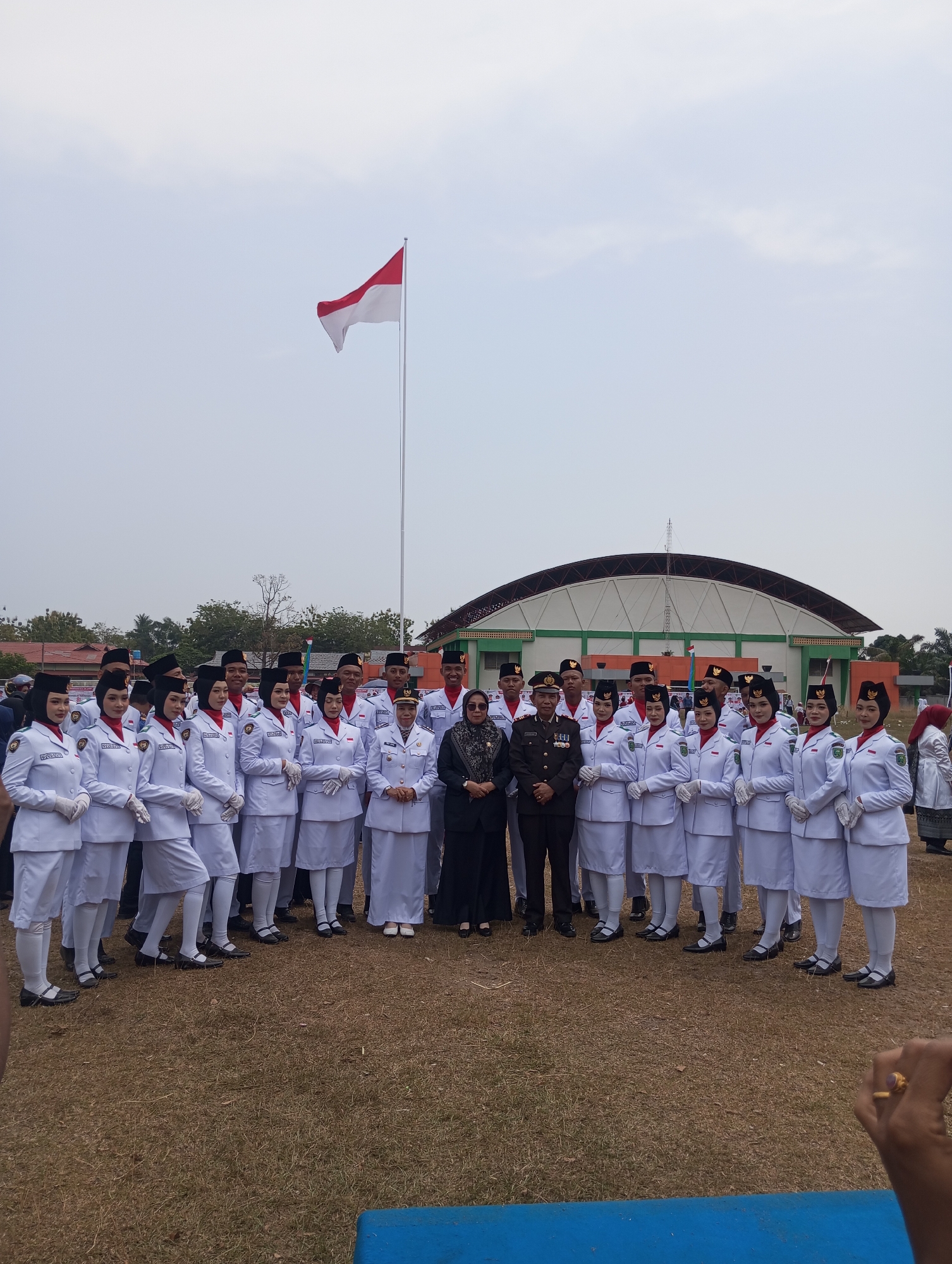 An outdoor group portrait shows a contingent of uniformed individuals, likely students or cadets, posing for a photograph. They are arranged in rows on a grassy field in front of a large, modern building with a curved roof, possibly a gymnasium or performance hall. The building is painted white with green and orange accents. A tall flagpole with the Indonesian flag (red and white) flying at the top stands to the right of the group, behind the individuals.
The uniformed individuals are mostly young women dressed in white blazers with red collars and white skirts or trousers. They are also wearing white gloves and black shoes with white socks, and many have black hats. A few young men in similar white uniforms and black hats are interspersed within the group. In the center front of the group, two individuals stand out: a man in a dark military-style uniform with medals, and a woman in a dark suit with a patterned headscarf. To the far left of the uniformed group, two women in dark clothing and headscarves are also present, seemingly observing. The overall atmosphere appears to be formal, possibly related to a ceremony or event. The sky is overcast and the ground is covered in dry grass. The scene is likely situated in Pematang Reba, Indonesia, given the national flag prominently displayed.