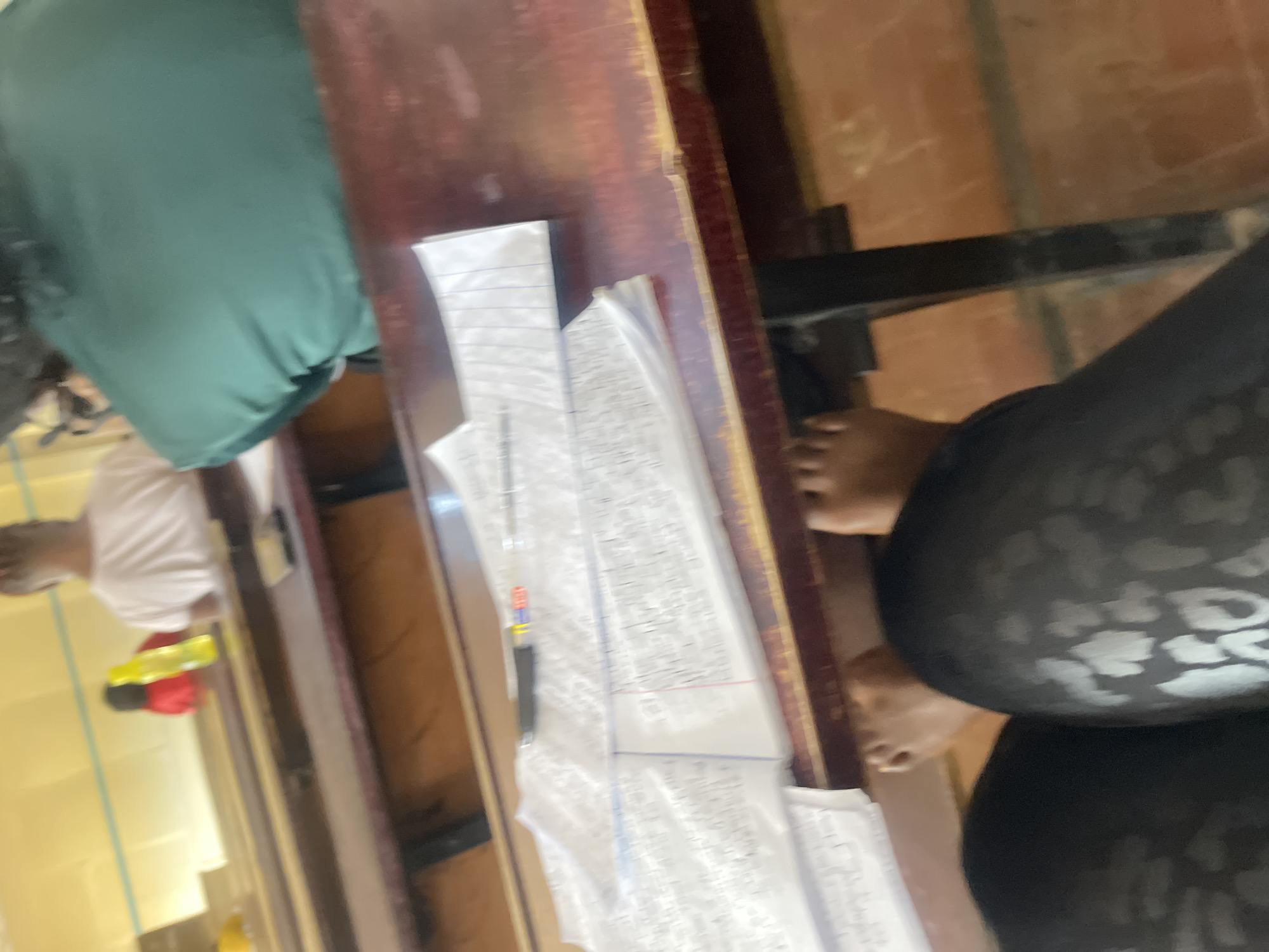 The image shows a classroom setting, likely in Rafin Par, Nigeria. In the foreground, a person's legs wearing black patterned leggings are visible, resting on a wooden desk. The desk surface is cluttered with papers and a pen. In the background, two other individuals are seated at desks. One person is wearing a white t-shirt, and another is wearing a green top. There is a yellow water bottle on a desk in the background. The overall environment appears to be a typical classroom with wooden desks and chairs. The lighting suggests it is daytime.