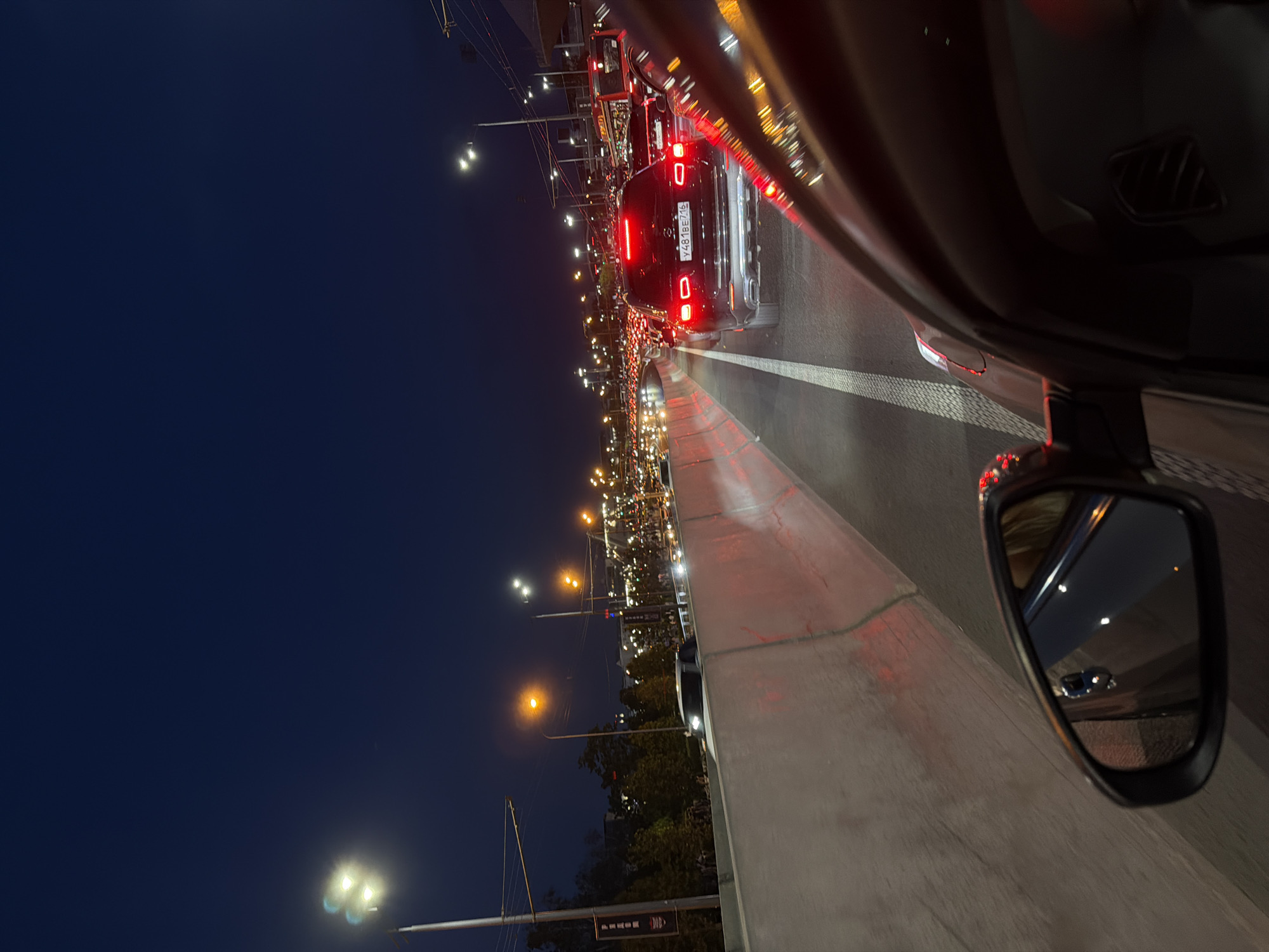 The image depicts a night scene of heavy traffic on what appears to be a bridge or overpass in Kazan, Russia. This is evident from the vehicle's license plate number Y481BE716, which is a Russian license plate number. Several cars are lined up ahead, with illuminated taillights. Street lights line the road and the dark night sky is visible in the background. The photograph is taken from inside a vehicle, looking out past the side mirror.
