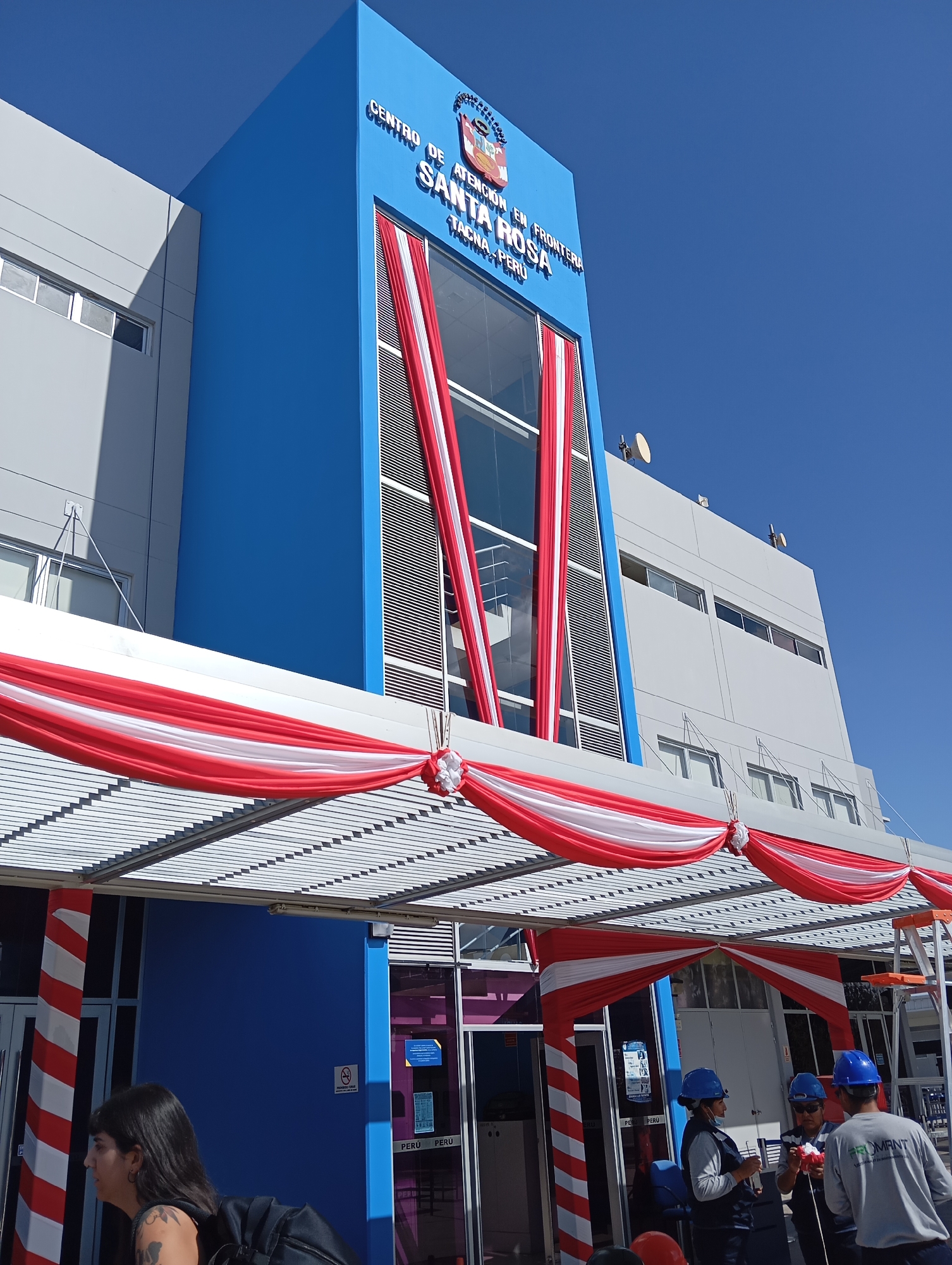 The image shows the exterior of the "Centro de Atención en Frontera Santa Rosa" in Tacna, Peru. The building features a prominent blue section with white lettering that reads "CENTRO DE ATENCIÓN EN FRONTERA SANTA ROSA TACNA - PERÚ". Red and white draped fabric decorations are hung across the entrance and along the facade of the building. Several individuals, some wearing hard hats, are visible near the entrance, possibly involved in setup or maintenance. The sky is clear and blue.