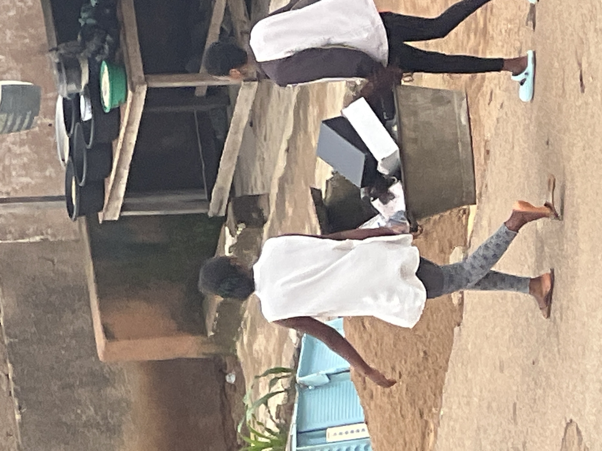 Two young individuals are walking away from the camera on a dirt path in what appears to be an outdoor market setting in the city of Jos, Nigeria. The individual on the left is wearing a white sleeveless shirt and grey patterned leggings, with brown sandals. The individual on the right is wearing a dark long-sleeved shirt with white panels on the sleeves and front, and black leggings with light blue slip-on shoes. They are walking past a large grey bin filled with various objects, including black and white boxes. In the background, a wooden table holds several black buckets and a green bowl. The surroundings are dusty and appear to be part of a developing urban area.