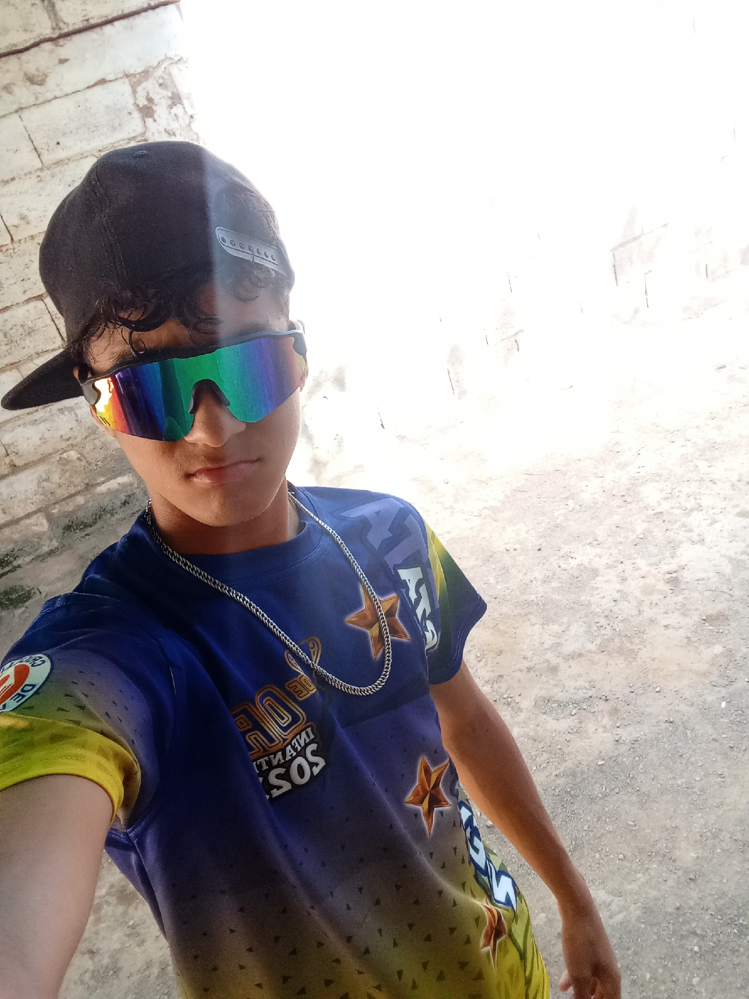 A young person stands in a brightly lit outdoor location in Juan Griego, Venezuela. They are wearing a black baseball cap tilted slightly back, iridescent sunglasses, a blue T-shirt with yellow sleeves and graphics, and a silver chain necklace. The background appears to be a concrete wall or building exterior on one side, and a brighter open space on the other. The ground is uneven and appears to be dirt or gravel.