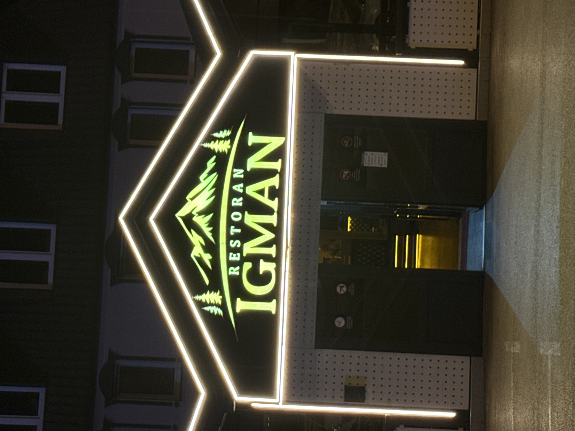 A neon sign displaying "RESTORAN IGMAN" is illuminated above the entrance to a building. The sign features a green mountain graphic with pine trees. The entrance itself has two large black doors. The building is lit with white neon lights forming a decorative outline around the sign and entrance. The ground in front of the building appears wet, reflecting the lights. The overall scene depicts the exterior of a restaurant at night.