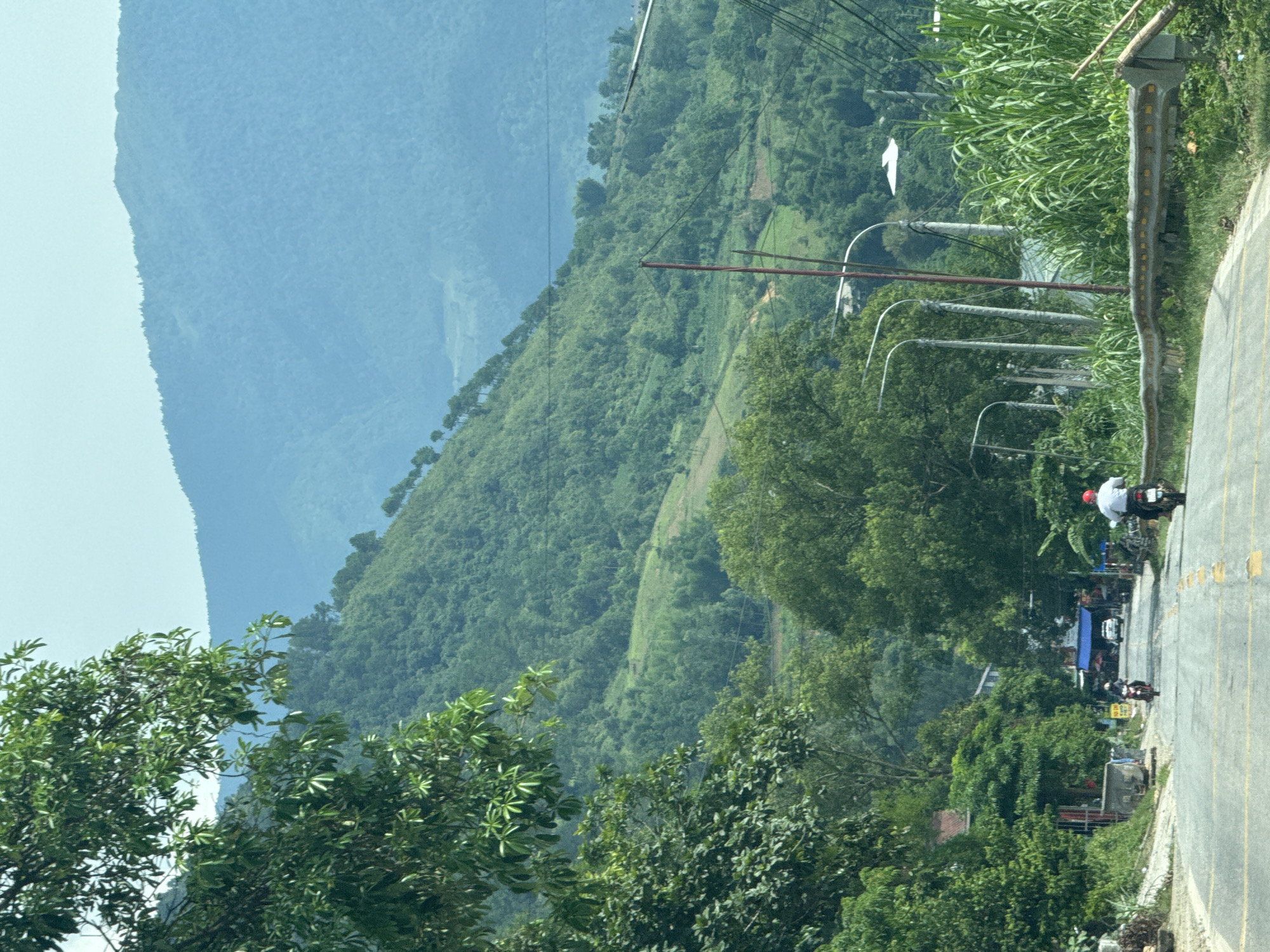 This image captures a vibrant, mountainous landscape in Muong Kim, Vietnam, bathed in bright daylight under clear skies. The scene is dominated by a succession of verdant, tree-covered mountains that extend into the horizon, with the most distant peaks appearing in soft, hazy shades of blue due to atmospheric perspective.
In the mid-ground and foreground on the right, a paved road with a yellow centerline (appearing as a double line due to perspective) runs along the side of a mountain, leading into the distance. A grey guardrail, marked with reflective yellow dots, flanks the road's edge. Along the roadside, dense green foliage, including tall grasses and numerous trees, frames the view.
Human presence is marked by infrastructure and individuals. Several utility poles with overhead power lines are visible along the road. On the road itself, a few motorcycles are traveling. One rider, distinctly wearing a white shirt and a red helmet, is seen on a white motorcycle, heading away from the viewer. Further along the road, another person on a dark motorcycle is also visible. In the distance, near the road, hints of human settlement or activity can be seen, including a blue awning that might belong to a small shop or market stall.
The image suggests a typical day in a scenic, rural mountainous area. No distinct text is clearly legible on any signs or objects within the frame. The overall impression is one of travel through a lush, green, and serene natural environment.