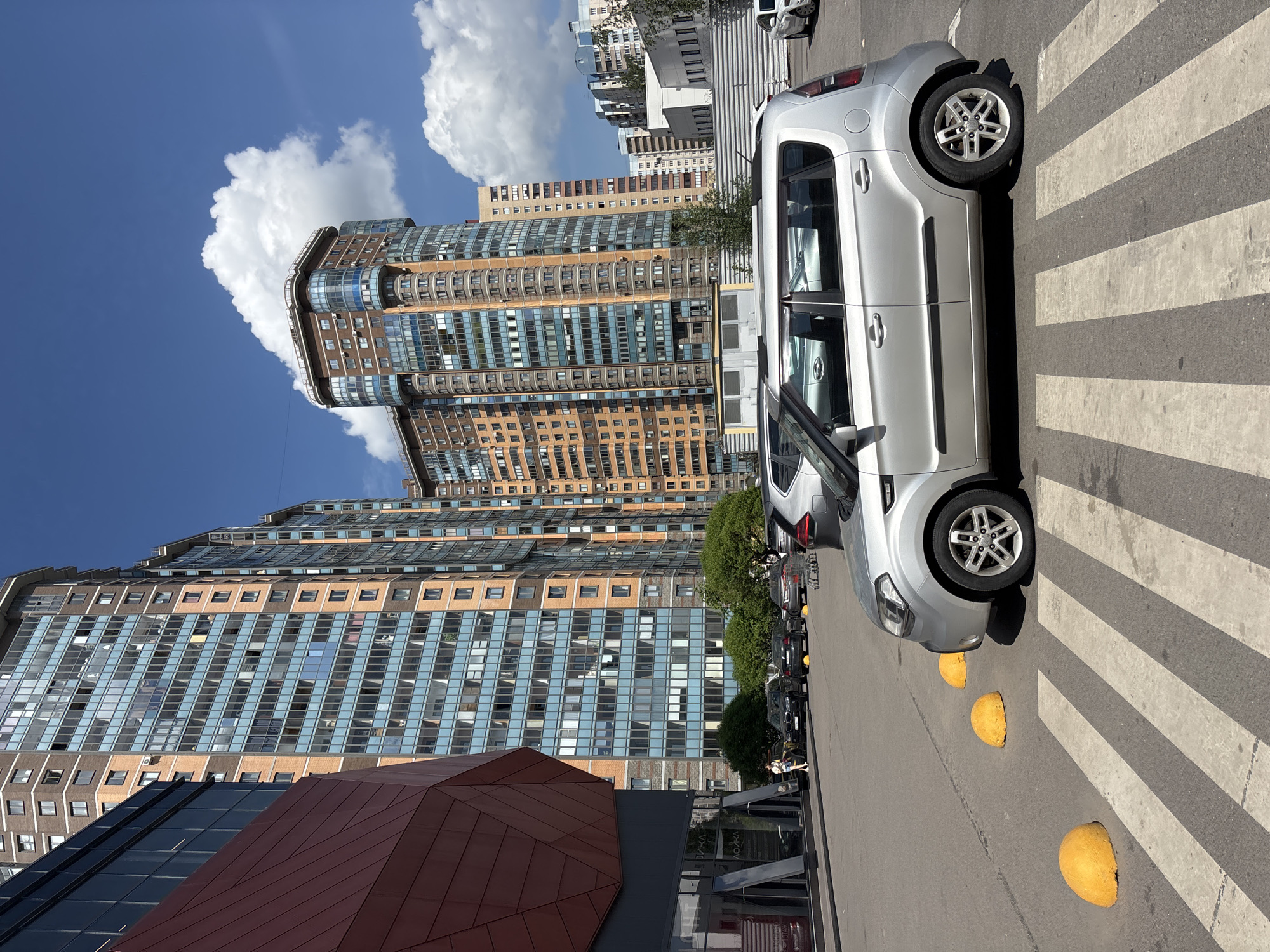 An urban scene unfolds under a bright blue sky, showcasing a blend of modern architecture and street infrastructure on a sunny day. In the foreground, a silver Kia Soul is prominently parked on a white-striped pedestrian crossing, facing to the right. To its immediate right, three yellow, hemispherical concrete barriers are spaced along the edge of the asphalt road. Behind the Kia, a line of other dark-colored cars is parked along the curb, partially obscured by green trees or bushes.
The background is dominated by several large, multi-story buildings. A distinctive high-rise in the mid-ground features a combination of tan-colored panels and extensive blue-tinted glass facades with curved sections and balconies. Another tall building further back and to the left appears to be constructed with brown or tan brick-like paneling and numerous windows. In the lower-left foreground, a modern building with a reddish-brown, angled metallic roof or facade is visible.
The sky is a vibrant blue, dotted with large, fluffy white cumulus clouds, suggesting clear and pleasant weather. The bright sunlight casts distinct shadows from the car and buildings. No people are visible, and no particular activity is taking place beyond the static urban setting. A subtle but significant detail is the visible text "ВЫПИВКА" (meaning "DRINKS" or "ALCOHOL" in Russian) on a storefront sign in the lower right background, providing a strong geographical cue for the image's location in Russia.