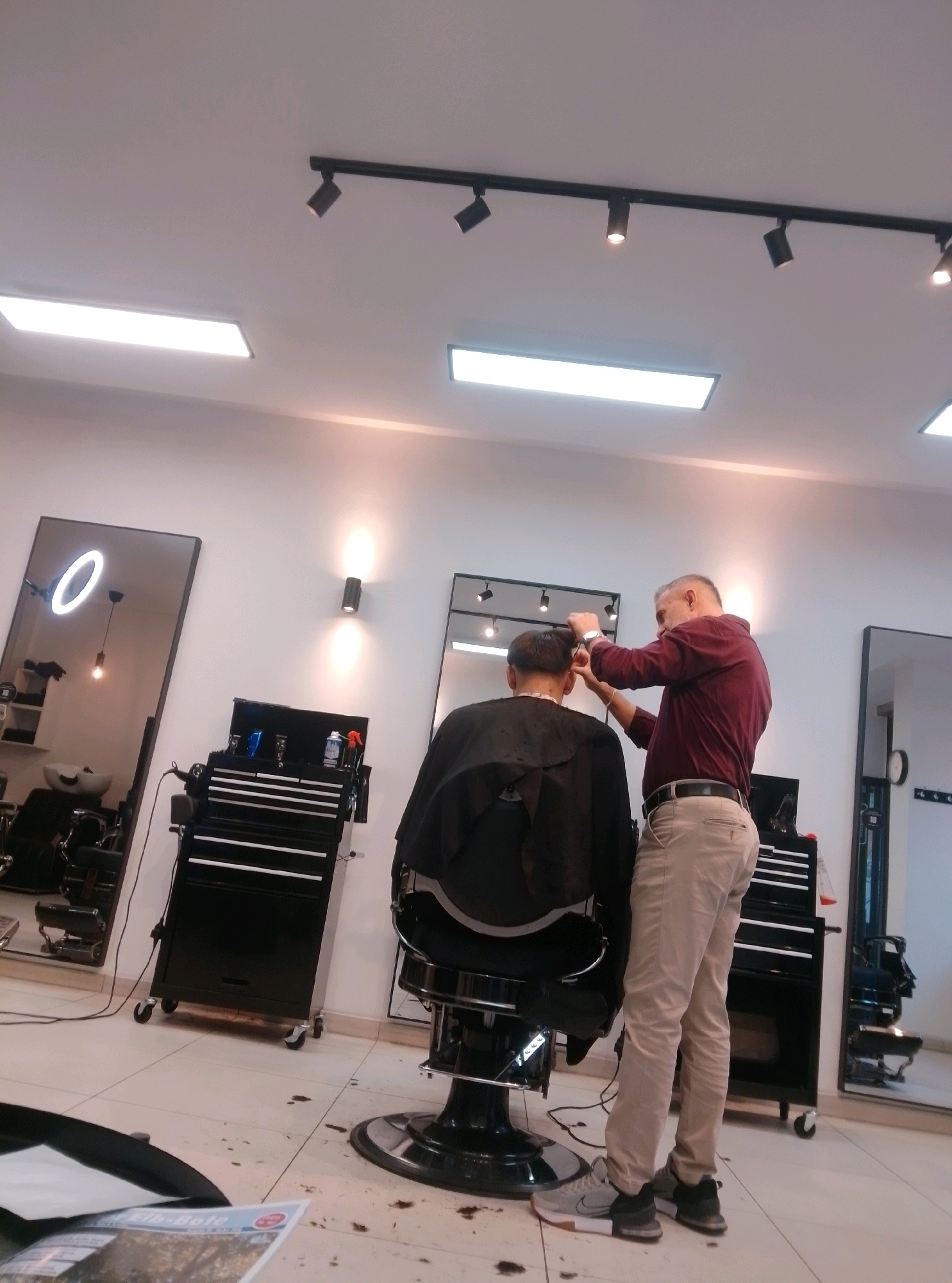 The image depicts an indoor scene, likely a barbershop, with two individuals. A man with graying hair, wearing a maroon long-sleeved shirt and light-colored trousers, stands behind another person seated in a black barber chair. The seated person, wearing a black cape, is partially visible from the back. The standing man appears to be in the process of cutting the seated person's hair, holding what looks like clippers or scissors in his right hand.
The environment features white walls and a tiled floor. Several black rolling tool cabinets are visible, one positioned near the barber chair and another further to the right. Large mirrors with illuminated ring lights are present on the left and partially visible on the right. Track lighting and wall-mounted fixtures illuminate the space. The overall impression is of a functional and modern barbershop. The presence of a magazine or brochure in the foreground suggests a customer waiting area or a display.
While the image is of a barbershop and shows a haircut in progress, there are no explicit visual cues within the image that specifically place the location in Wedel, Germany. The description is limited to the observable elements in the photograph.