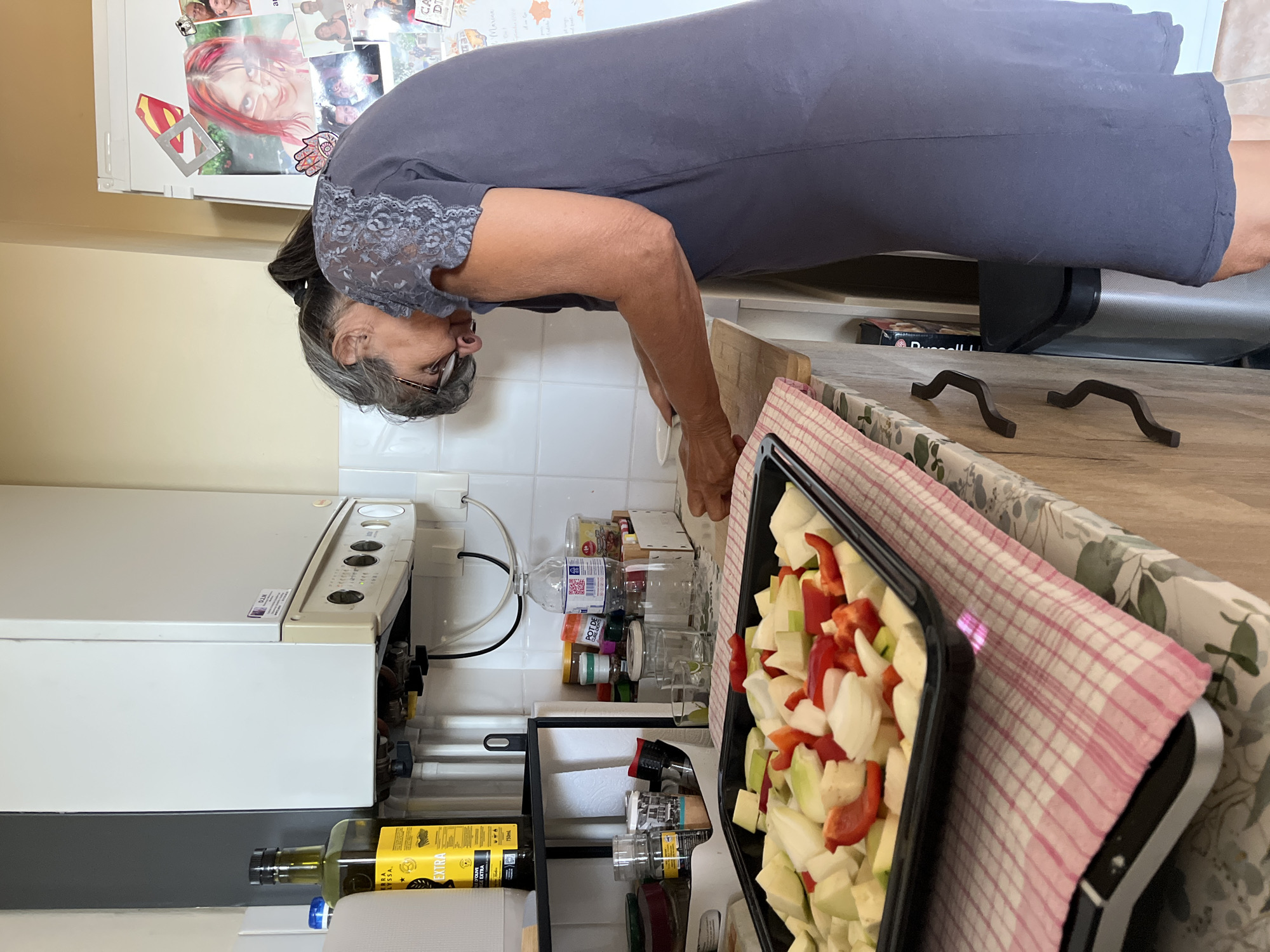 A middle-aged woman with graying hair pulled back in a ponytail is standing in a kitchen, leaning over a countertop. She is wearing a dark gray, sleeveless dress with lace detailing on the shoulders. Her arms are extended as she appears to be preparing food. In front of her is a black baking dish filled with chopped vegetables, including what appear to be bell peppers, zucchini, and onions. The baking dish rests on a pink and white checkered cloth, which is placed on a countertop with a floral pattern. To the left of the baking dish is a bottle of olive oil. In the background, a white boiler is mounted on the wall, and a refrigerator is visible on the right, adorned with various photographs and notes. The kitchen appears to be in a residential setting, possibly an apartment or house, and given the context, it is likely located in Montélimar, France.