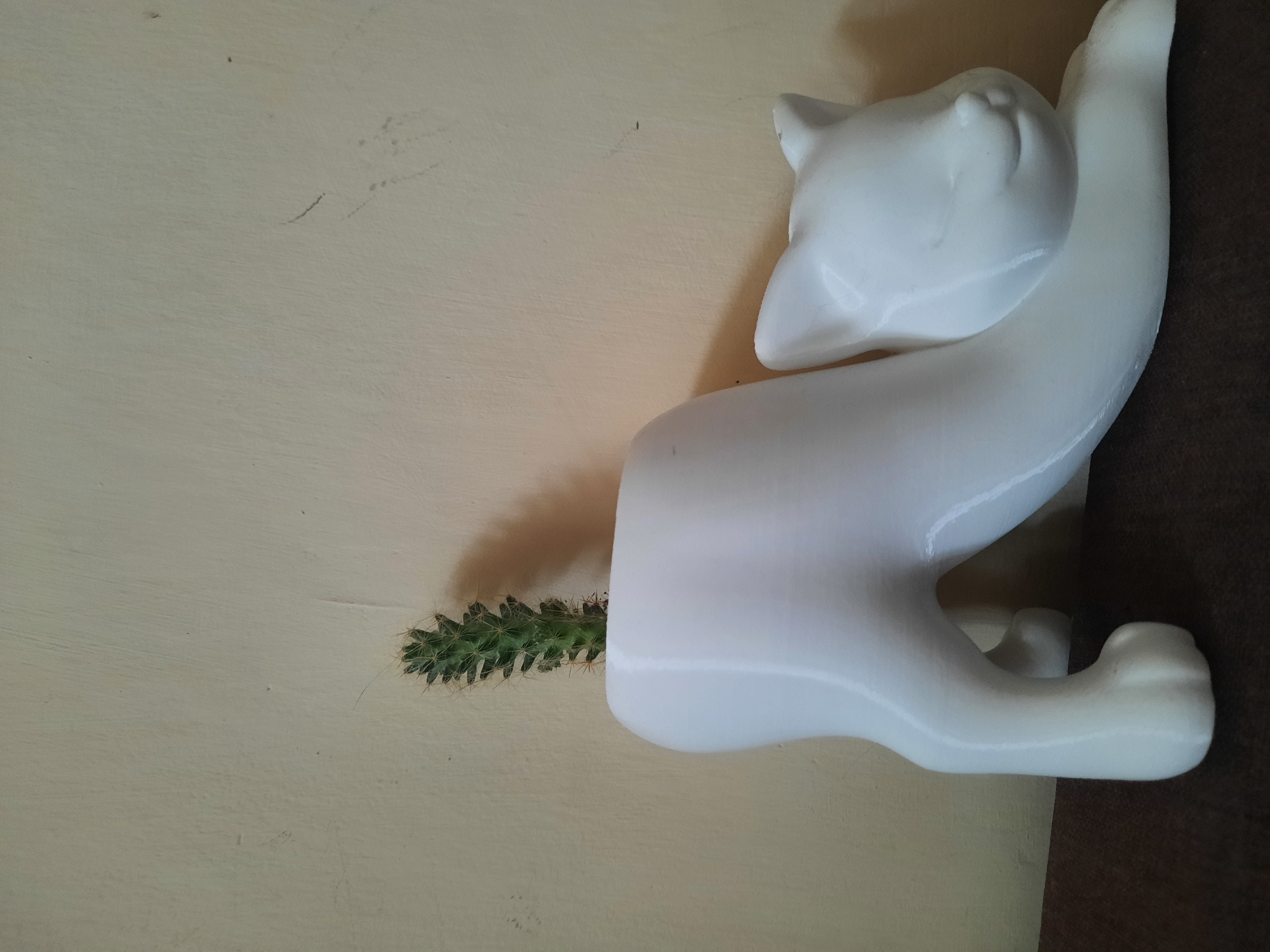 A white ceramic planter, shaped like a reclining cat, holds a small green cactus. The planter is placed on a dark brown surface against a plain, light-yellow wall. The cat's head is turned to the right, with closed eyes and a subtle smile. Its body is stretched out, with its hind legs tucked underneath and its front paws extended forward. The cactus, positioned in the cat's back, is narrow and spiky. The scene appears to be indoors, possibly on a table or shelf. The lighting suggests ambient room light, casting a soft shadow behind the planter on the wall. There is no visible indication of the city of Blagodarny, Russia, in this image.
