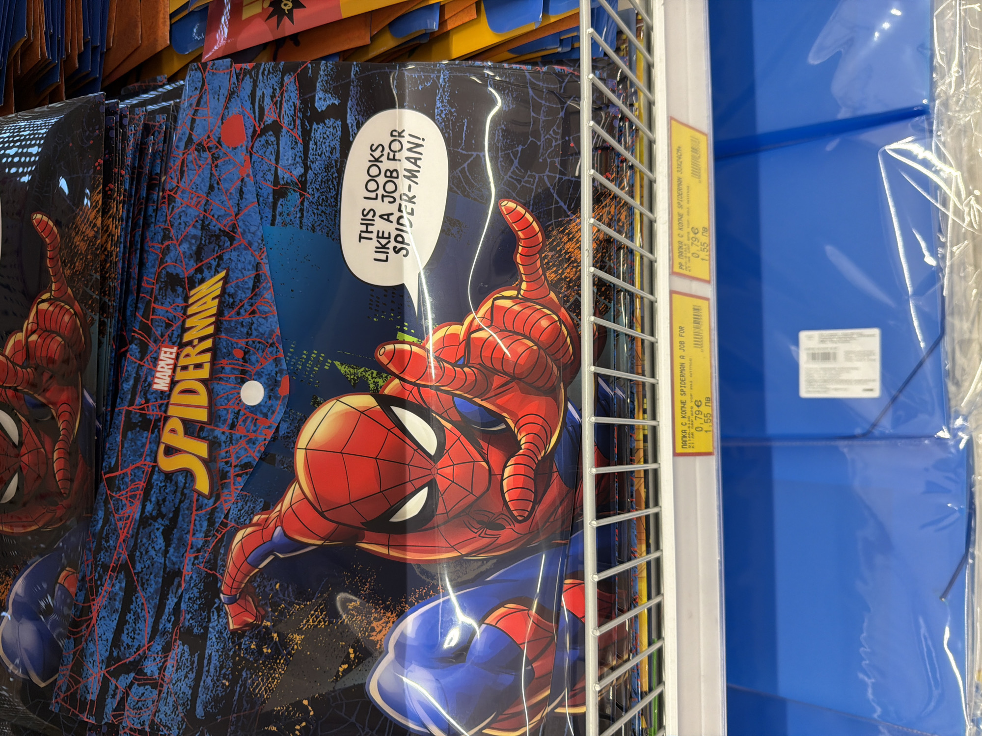 The image shows a close-up of a display shelf in a store, likely a stationery or toy shop, in Plovdiv, Bulgaria. The main subject is a stack of folders or portfolios featuring the Marvel character Spider-Man. The designs include the Spider-Man logo, Spider-Man in action poses, and a speech bubble with the text "THIS LOOKS LIKE A JOB FOR SPIDER-MAN!". The folders are packaged in plastic. Below the Spider-Man items, there are blue folders. Two yellow price tags are visible. One tag, for "PP папка с копче SPIDERMAN 33x24CM", indicates a price of "0,79 € / 1,55 лв". The other tag, partially obscured, also mentions "SPIDERMAN". The environment appears to be indoors with fluorescent lighting. The overall scene depicts merchandise for sale in a retail setting.