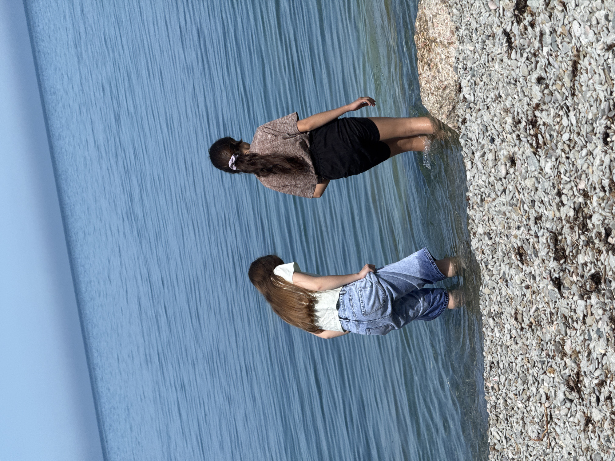 Two young women are standing in shallow water at the edge of a beach. The woman closest to the viewer is wearing a white shirt, denim shorts, and has long brown hair. The other woman is wearing a brown shirt, black shorts, and has long brown hair tied back with a white scrunchie. The beach is covered in small pebbles. The water is calm and blue. The location is likely a beach, but the city is unknown.