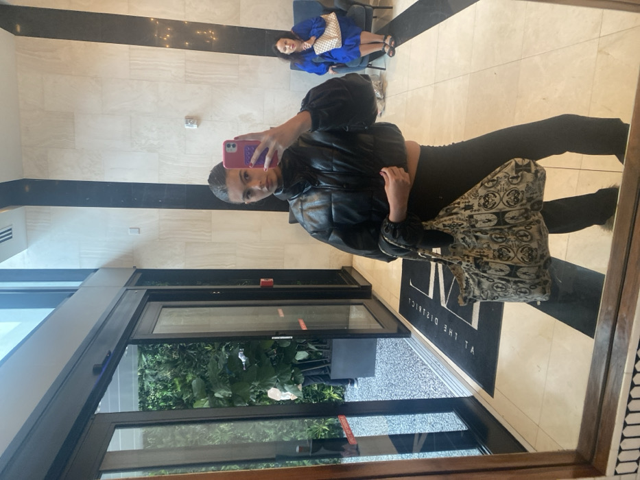 A reflection in a mirror shows a young woman taking a selfie. She wears a black puffer jacket, black pants, and a patterned handbag. Behind her is a well-lit lobby with neutral-toned tiled walls and a black accent stripe. A doorway is visible, showing greenery outside. In the distance, a person in a blue outfit sits on a chair. The floor is light-colored and reflects the scene. "AT THE DISTRICT" is imprinted on a dark doormat.