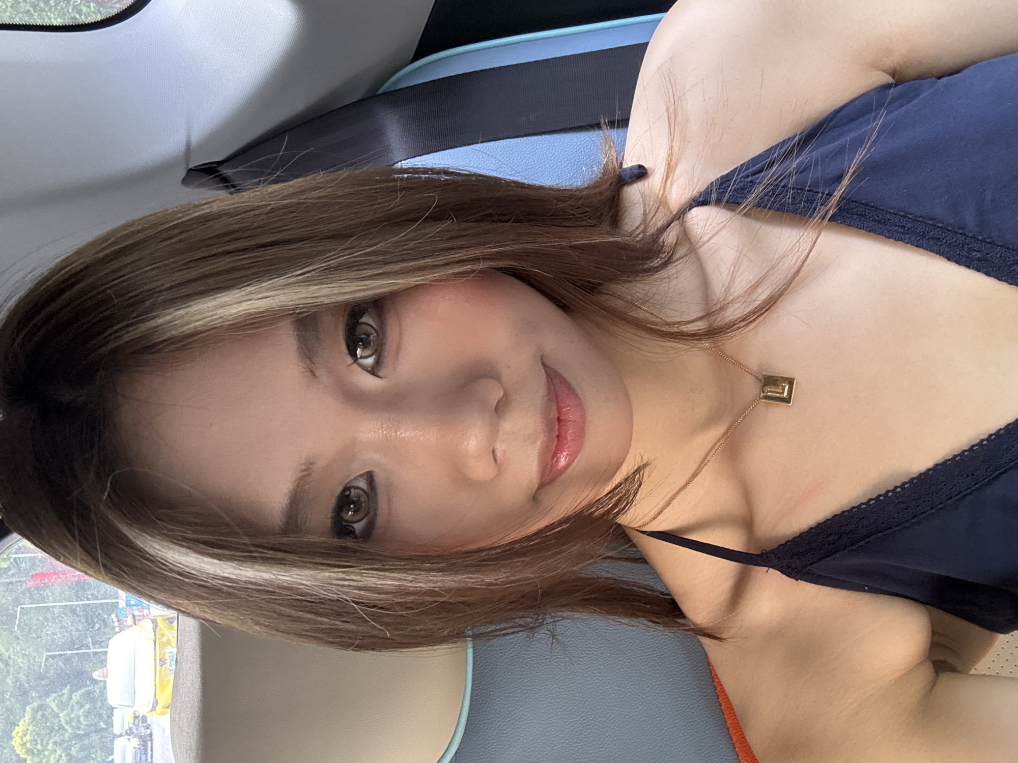 A medium shot depicts a woman inside a vehicle, possibly a car. She has brown hair with blonde highlights and wears a dark blue tank top with a lace trim. A gold necklace with a rectangular pendant adorns her neck. A seat belt is visible behind her. The interior of the vehicle is light gray and blue. Outside the vehicle's window, there are trees and various structures. While the photograph's caption specifies the location as Nan'an District, China, this cannot be confirmed directly from the visual elements in the image.