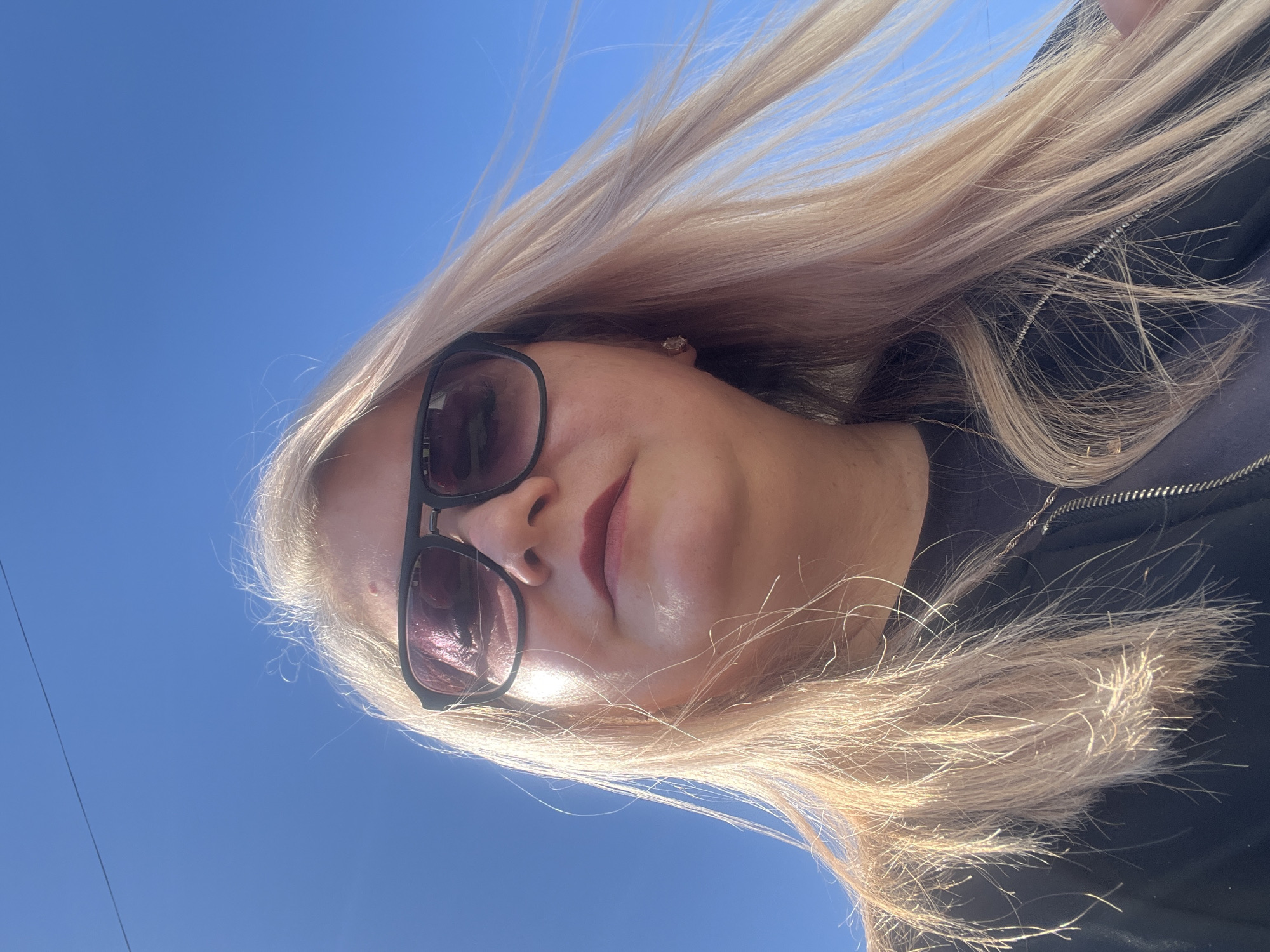 A woman with long blonde hair is photographed against a clear blue sky. She is wearing sunglasses and a dark jacket. The image appears to be taken from a low angle, looking up at the subject. There is no information in the image that indicates the location is Glazov, Russia.