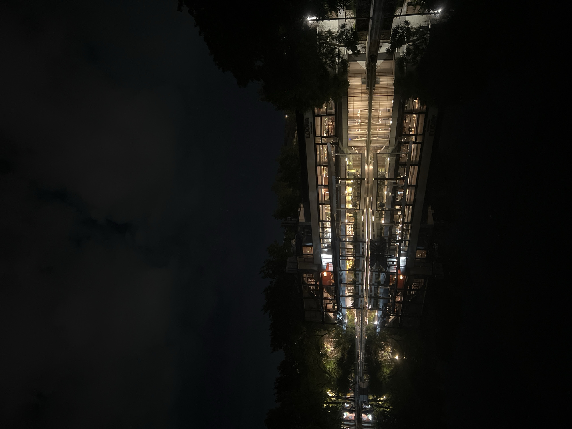 This media file captures a modern, multi-story building illuminated at night, perfectly reflected in a calm body of water. The scene is dominated by the building's contemporary architecture, featuring numerous glass panels, balconies, and warm interior lighting. Each floor of the structure appears active, with lights on, suggesting commercial or residential use.
Visible text on the building includes a sign that reads "LOONA Restaurant", indicating a dining establishment. Another sign, partially visible, displays the word "NUR". Dark foliage and trees are visible around the base of the building, providing a natural contrast to the man-made structure and also contributing to the reflection.
The time of day is clearly night, as evidenced by the dark sky, which shows faint cloud formations in the upper left. The stillness of the water creates a nearly flawless, symmetrical reflection of the building and its lights, making it appear as if the building extends downwards into an ethereal twin. The overall atmosphere is one of urban tranquility and sophistication, with the glowing lights and their reflections creating a captivating visual effect in the night setting of Yerevan, Armenia.