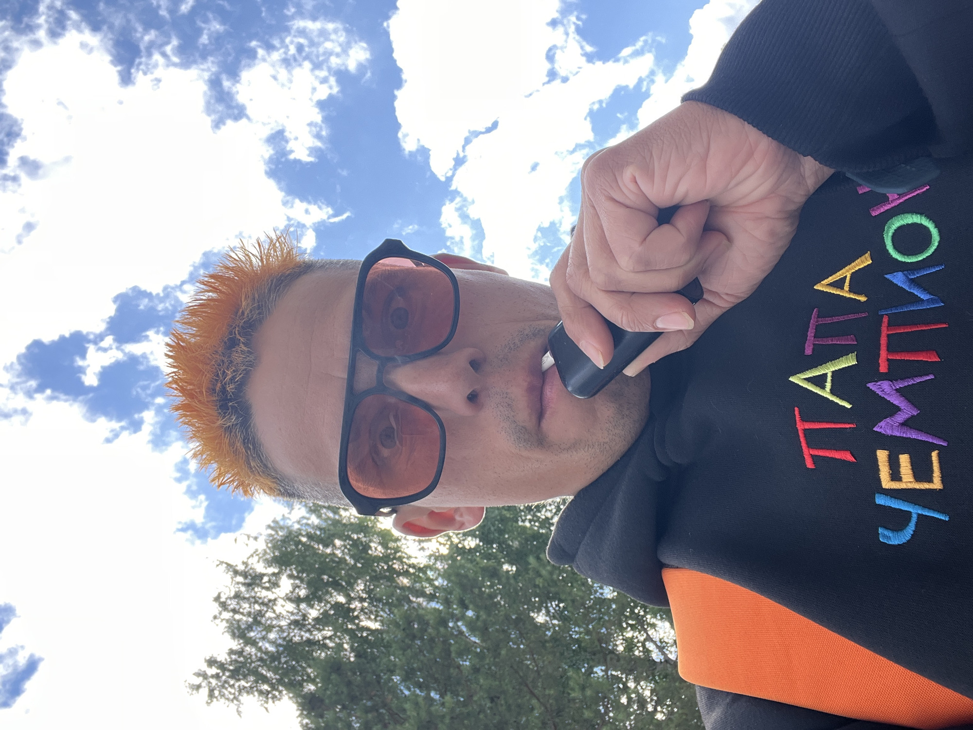 A low-angle, close-up shot features a person with vibrant orange hair, wearing sunglasses with orange-tinted lenses. They are holding a dark-colored object, possibly a vape pen, to their lips. The person is dressed in a black hoodie with colorful embroidered text on the right side. The text is a vertical arrangement of letters in various bright colors: pink, red, yellow, light green, blue, and purple. An orange garment is visible beneath the hoodie.
The background consists of a bright blue sky filled with fluffy white clouds. In the lower portion of the frame, green foliage from trees is visible, suggesting an outdoor setting. The lighting and cloud formations indicate it is daytime, likely a sunny day with scattered clouds. The upward angle of the shot, looking towards the sky, creates a dynamic perspective. The person's expression, partially obscured by sunglasses, is unreadable. No specific emotional cues are strongly discernible. No location-specific landmarks are visible, beyond the general context of an outdoor, possibly park-like or natural environment. The visible text is descriptive and seems to be a brand name or slogan.