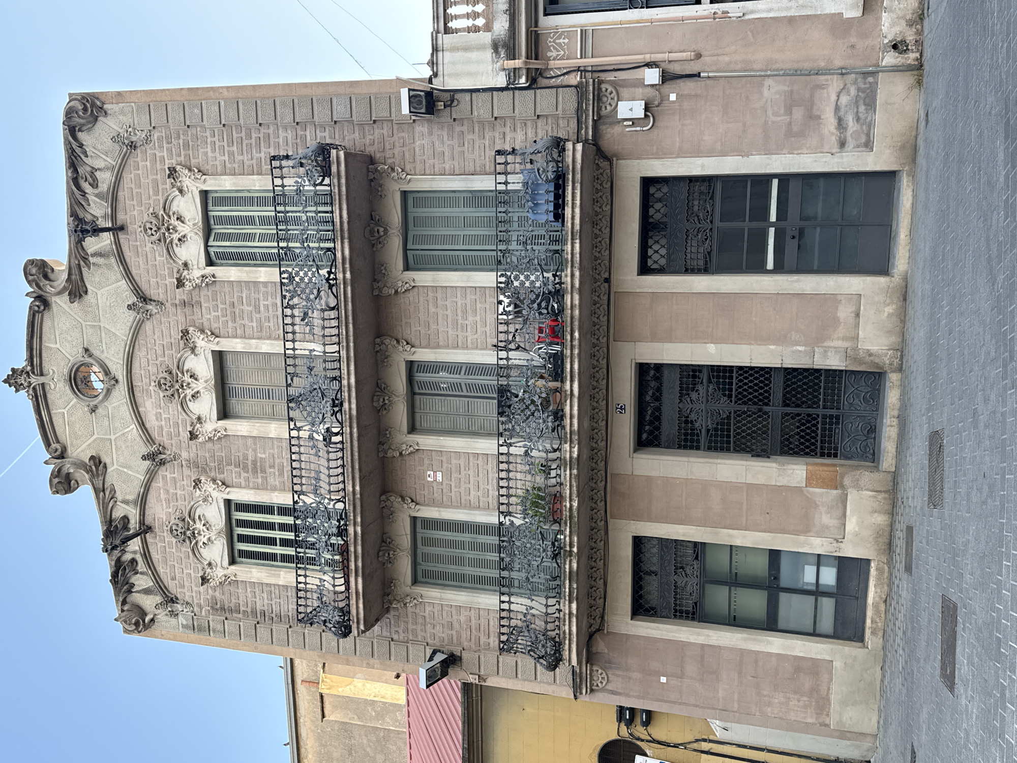 The image depicts a building with a distinctive architectural style located in Barcelona, Spain. The structure exhibits elements of Catalan Modernism, characterized by its ornate facade, decorative wrought-iron balconies, and intricate stonework.
The building has three stories, each featuring windows with shutters. The balconies, adorned with elaborate iron designs, are positioned on the second and third floors. The ground floor has barred windows.
The building's facade is decorated with carved stone details, particularly around the windows and roofline. The exterior is painted in a muted color scheme, highlighting the architectural details.
The location is a street in Barcelona, evident from the surrounding urban environment and architectural style. A sidewalk is visible.