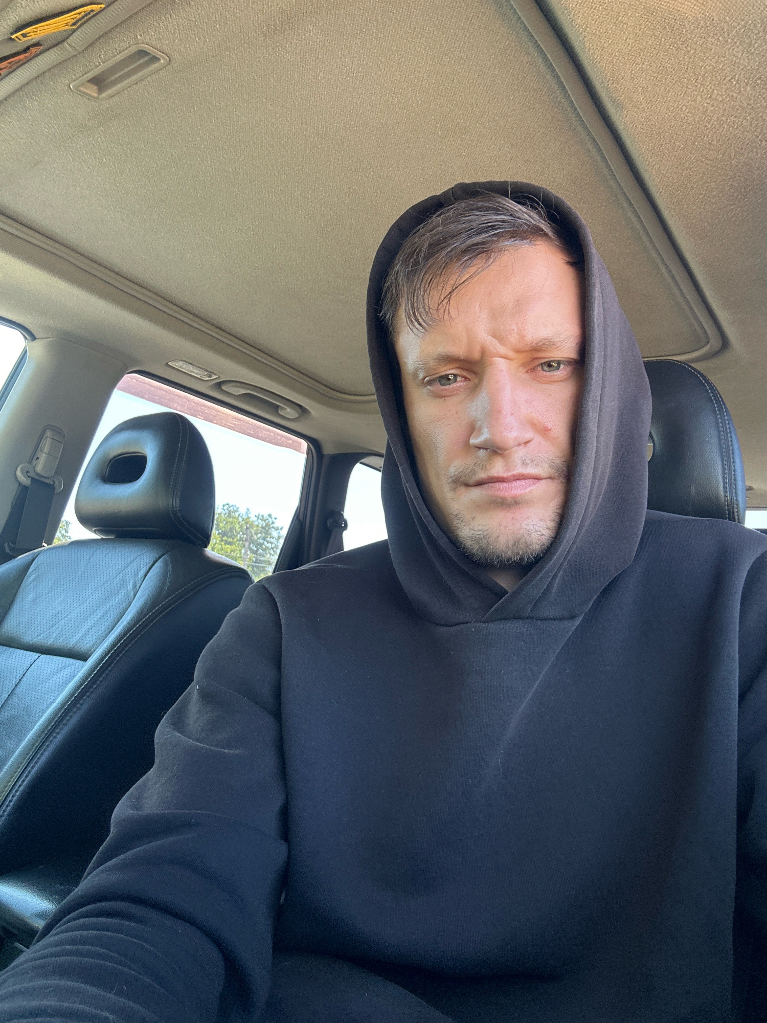 A medium shot selfie shows a man in a black hooded sweatshirt sitting in the driver's seat of a car. He is looking directly at the camera with a furrowed brow and a slight grimace. The man has light skin, brown hair, and stubble on his chin. The interior of the car is visible, including the black leather seats, the car's sunroof, and the car's headliner. Outside the car, trees and a blue sky are visible through the windows. The image appears to have been taken during the daytime. The location of Makiv, Ukraine, is not discernible from the image itself.