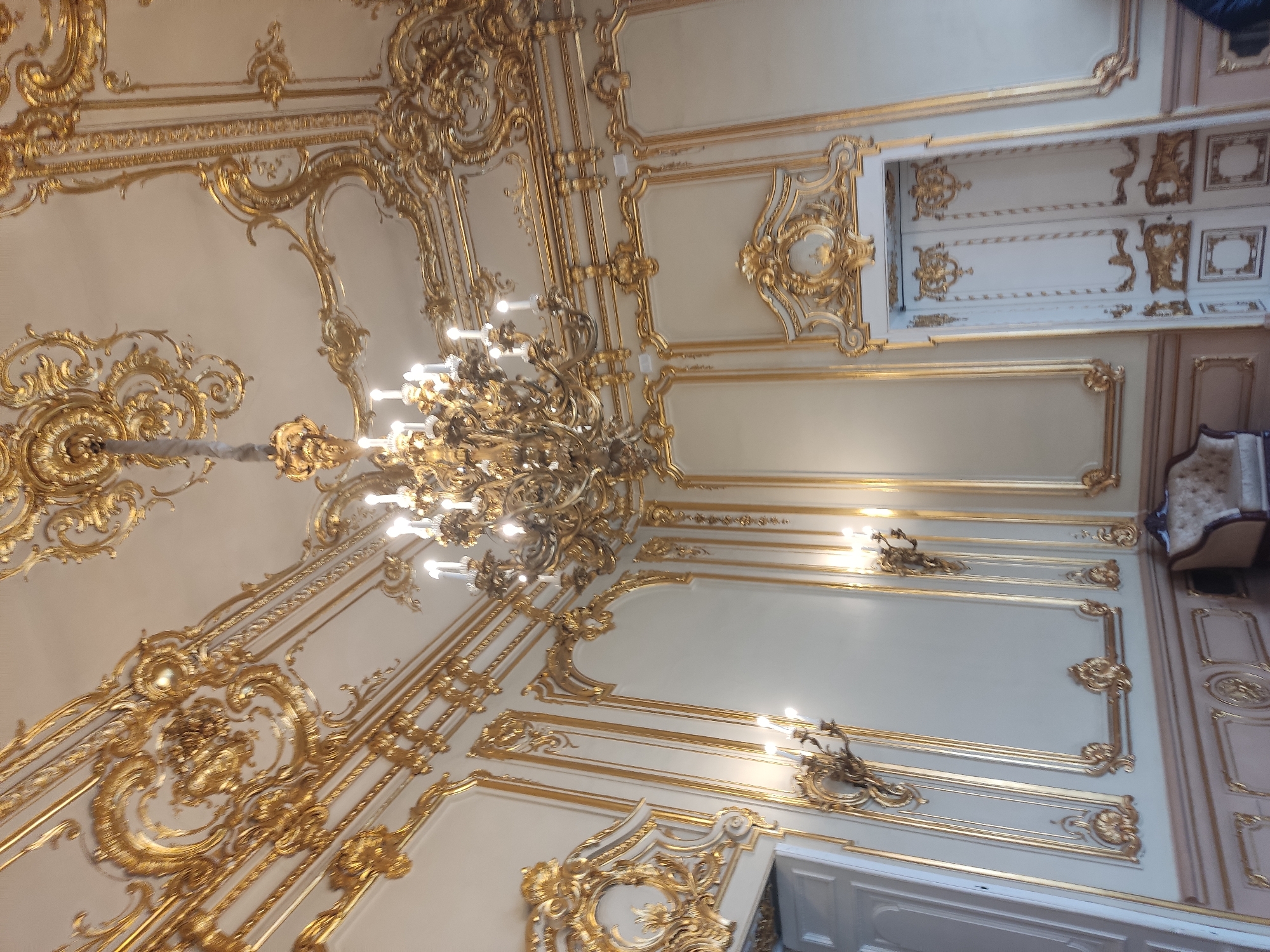 The image captures the opulent interior of a building in Saint Petersburg, Russia, characterized by elaborate, gilded decorations. A large, ornate chandelier dominates the frame, suspended from the ceiling with intricate gold detailing. The walls are embellished with gilded moldings, panels, and decorative flourishes. Architectural elements like doorways are adorned with ornate gold trim. The space appears well-lit, highlighting the metallic sheen of the gold embellishments. A glimpse of furniture, including an upholstered chair or sofa, can be seen in the background. The scene suggests a historical, luxurious interior, likely a palace or grand building within Saint Petersburg.