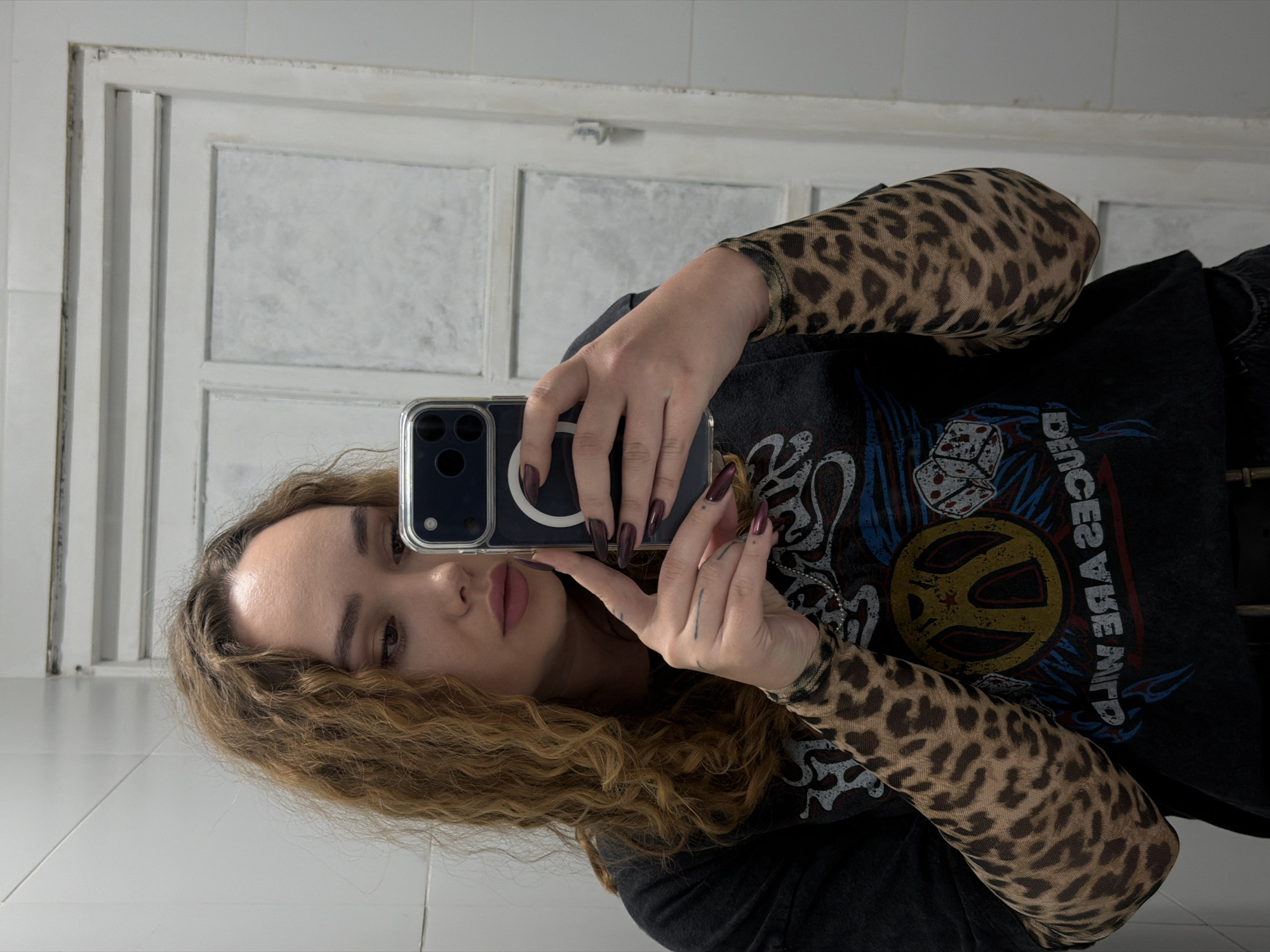 A young woman is taking a selfie in a bathroom. She is holding a smartphone in her right hand and is facing a mirror. She has long, curly brown hair and is wearing a dark t-shirt over a leopard-print long-sleeve shirt. She also has long, dark red fingernails. The bathroom has white tiled walls and a white door. There is no indication of the city or location being Ramat Gan, Israel.