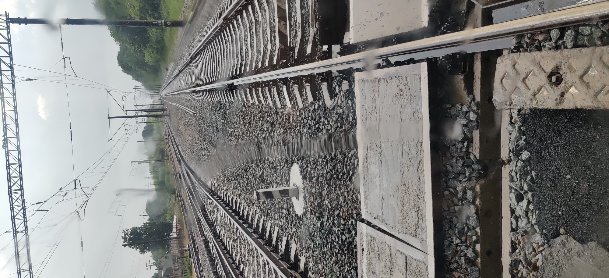 The image depicts a view of multiple train tracks, likely in a rural or suburban setting, given the presence of trees and a cloudy sky. The tracks appear to be part of a railway yard or junction, as evidenced by the converging and diverging lines of rails. Some of the tracks are covered in gravel ballast, while others have concrete sleepers. There are overhead electrical wires and poles visible, indicating an electrified railway line. The ground around the tracks is wet, suggesting it has recently rained or is currently raining, and there are puddles of water visible. A small, striped marker post is present near the center of the image. The overall scene is one of infrastructure for transportation in potentially inclement weather.
The specific location in the city of Shelestove, Ukraine, is not directly discernible from the visual information presented in the image. While the image shows railway tracks and associated infrastructure, there are no unique landmarks, signage, or contextual clues that would definitively place it in Shelestove or any other specific city. Therefore, any statement about the location being Shelestove, Ukraine, would be an assumption or external information not derivable from the photograph itself.