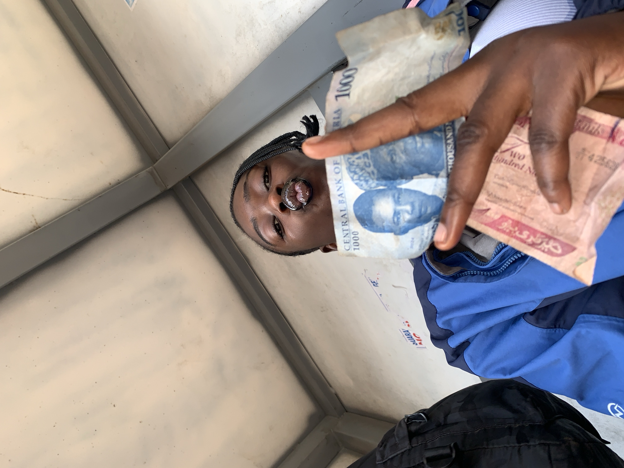 The image is a close-up, selfie-style shot of a young person holding Nigerian currency. The person is wearing a blue jacket and has their lips puckered, with a lip ring visible. They are holding up a 1000 Naira note, with another bill partially visible in their hand. The background appears to be an outdoor structure with a white, possibly metallic, ceiling and grey beams. There is a black backpack on the left side of the frame. The location is identified as Rafin Par, Nigeria.