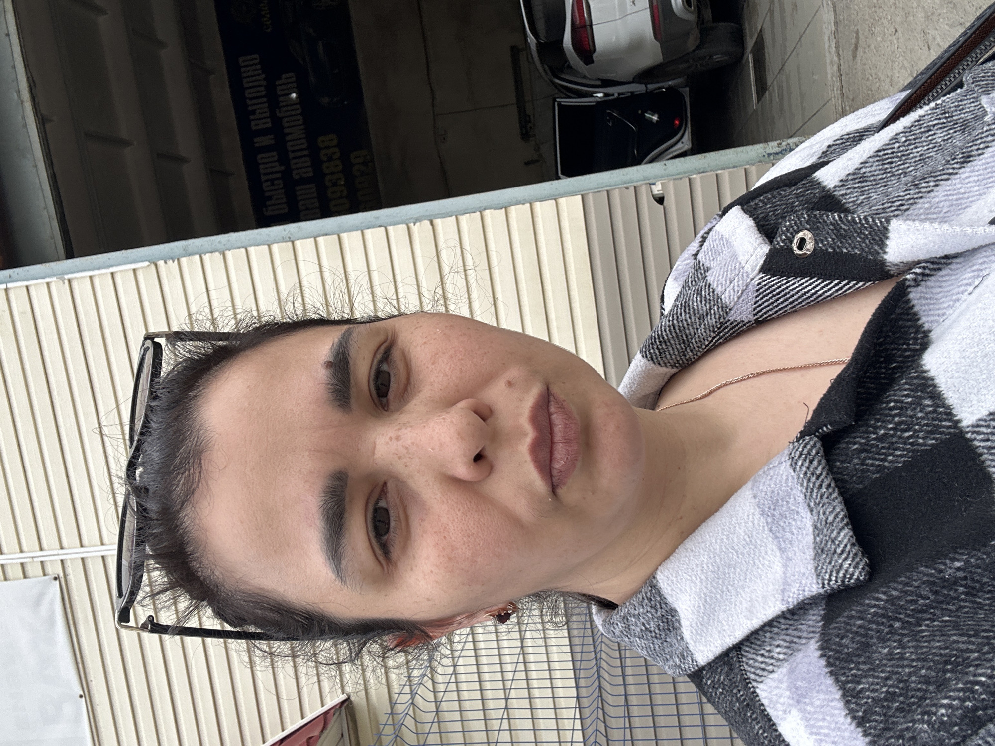 A rotated close-up portrait shows a woman, likely in her late 20s to 40s, looking directly at the camera with a neutral expression. She has dark hair pulled back, dark eyebrows, and visible pores on her skin. Glasses are perched on top of her head. She is wearing a black and white plaid shirt or jacket, and a thin gold-colored necklace is visible.
The background to her left features a light-colored, vertically ribbed wall or siding. To her right and behind her, a darker interior space is visible, containing the rear of a white car and parts of another dark vehicle. A dark sign with white, inverted Russian text is visible. The text reads: "БЫСТРО И ВЫГОДНО" (Quickly and profitably), followed by "ваш автомобиль" (your car), and numbers "093838" and "0029". This suggests the location is a car-related business, such as an auto repair shop or dealership. The lighting is soft and diffused, indicating an overcast day or indirect indoor illumination.
