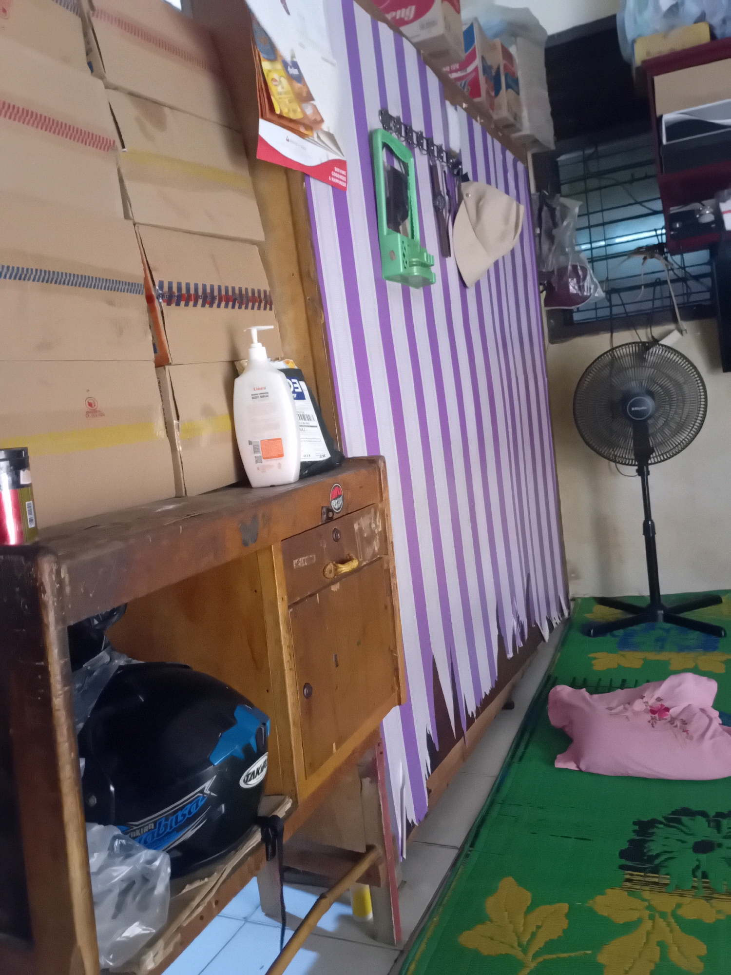 The image depicts an indoor scene, likely a room within a residential dwelling in Pekanbaru, Indonesia. On the left, stacks of cardboard boxes are visible, suggesting storage or a recent delivery. A wooden shelf unit holds various items, including a white pump bottle of lotion or soap, a black helmet with blue accents and a "TAKIBUSA" logo, and a red and silver cylindrical container.
Dominating the center and right of the image is a wall covered with purple and white vertical stripes, possibly wallpaper or a fabric covering. A light green wall-mounted organizer with hooks is attached to this striped wall, holding a beige cap. To the right of the striped wall, a black standing fan with a circular grille is positioned on a green mat with a floral pattern. A pink shirt is also laid out on the green mat. The overall impression is one of a cluttered living space or storage area in a home.