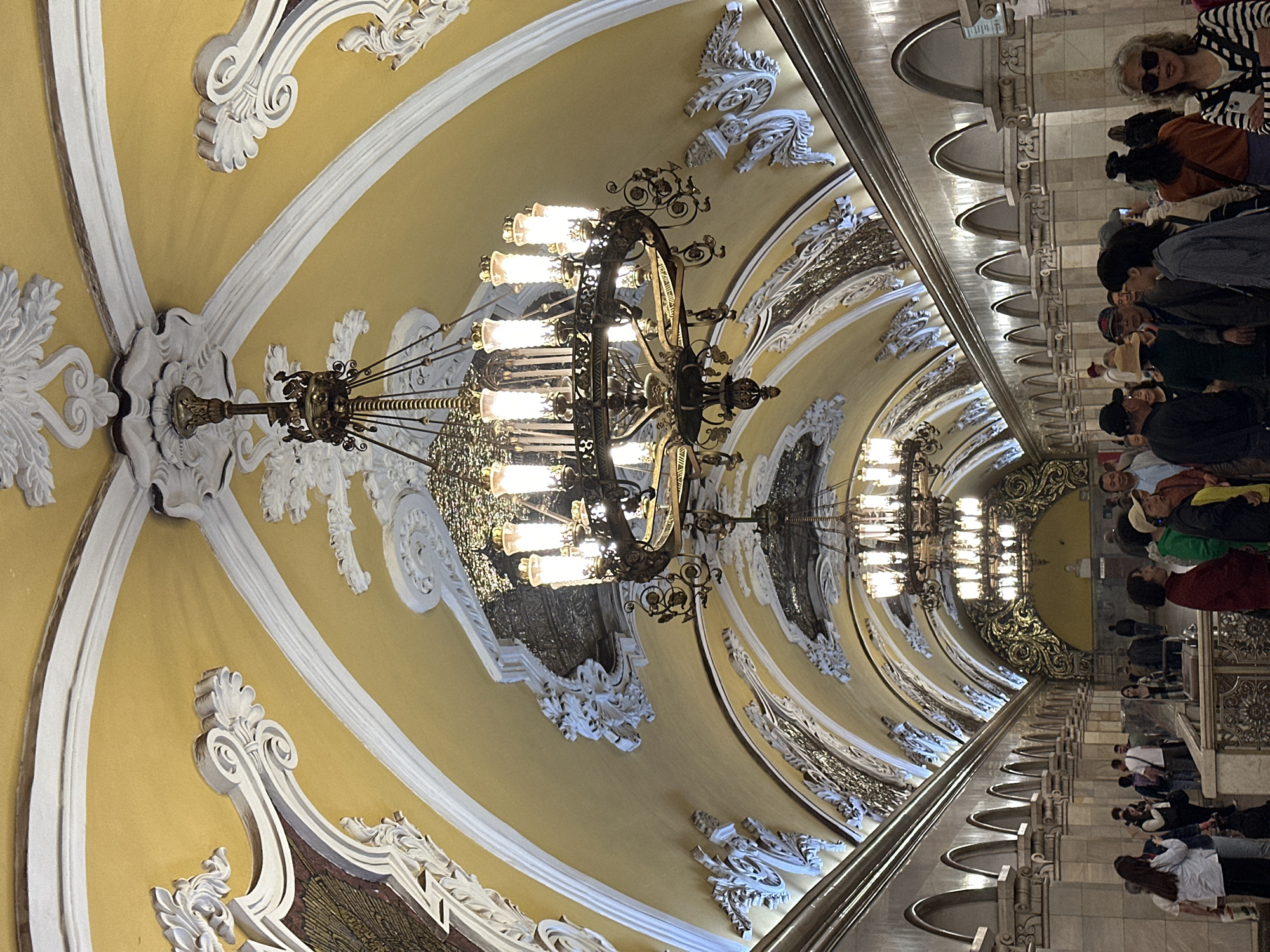 The image shows a section of the Komsomolskaya station on the Koltsevaya Line of the Moscow Metro. The architecture is Stalinist Baroque. Visible is the ornate yellow ceiling with white baroque-style decorations, and chandeliers. Many people are standing and walking along the platform.