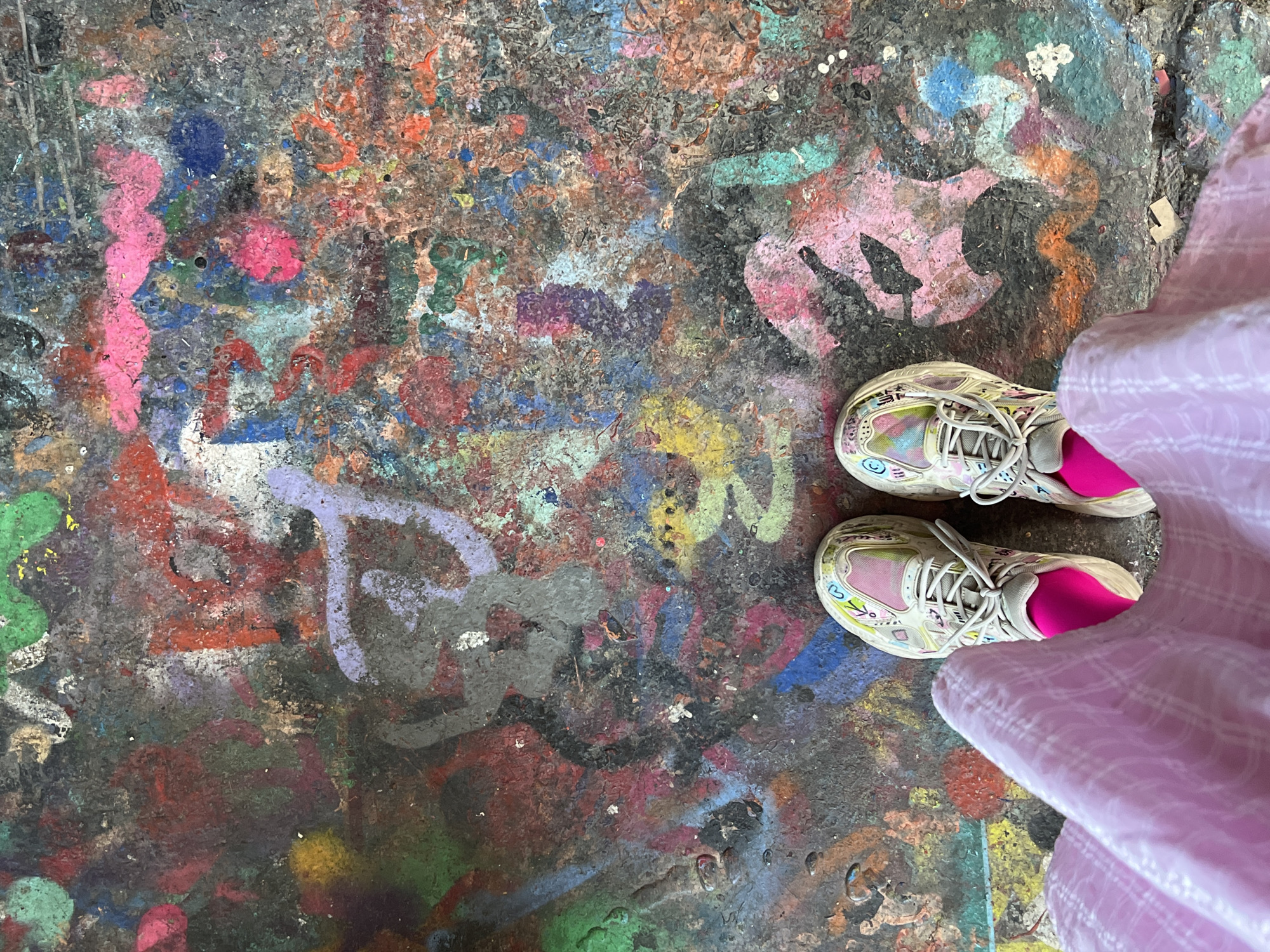 A top-down shot shows a person standing on an uneven, graffiti-covered surface. The person is wearing white sneakers with a pink sock liner and a light pink, possibly linen or cotton, plaid skirt or dress. The graffiti on the ground appears to be multi-layered and includes various colors such as pink, blue, green, yellow, and purple. The overall texture of the ground suggests it is aged and possibly concrete. The specific location is the "Beton Hala" graffiti wall in Belgrade, Serbia.