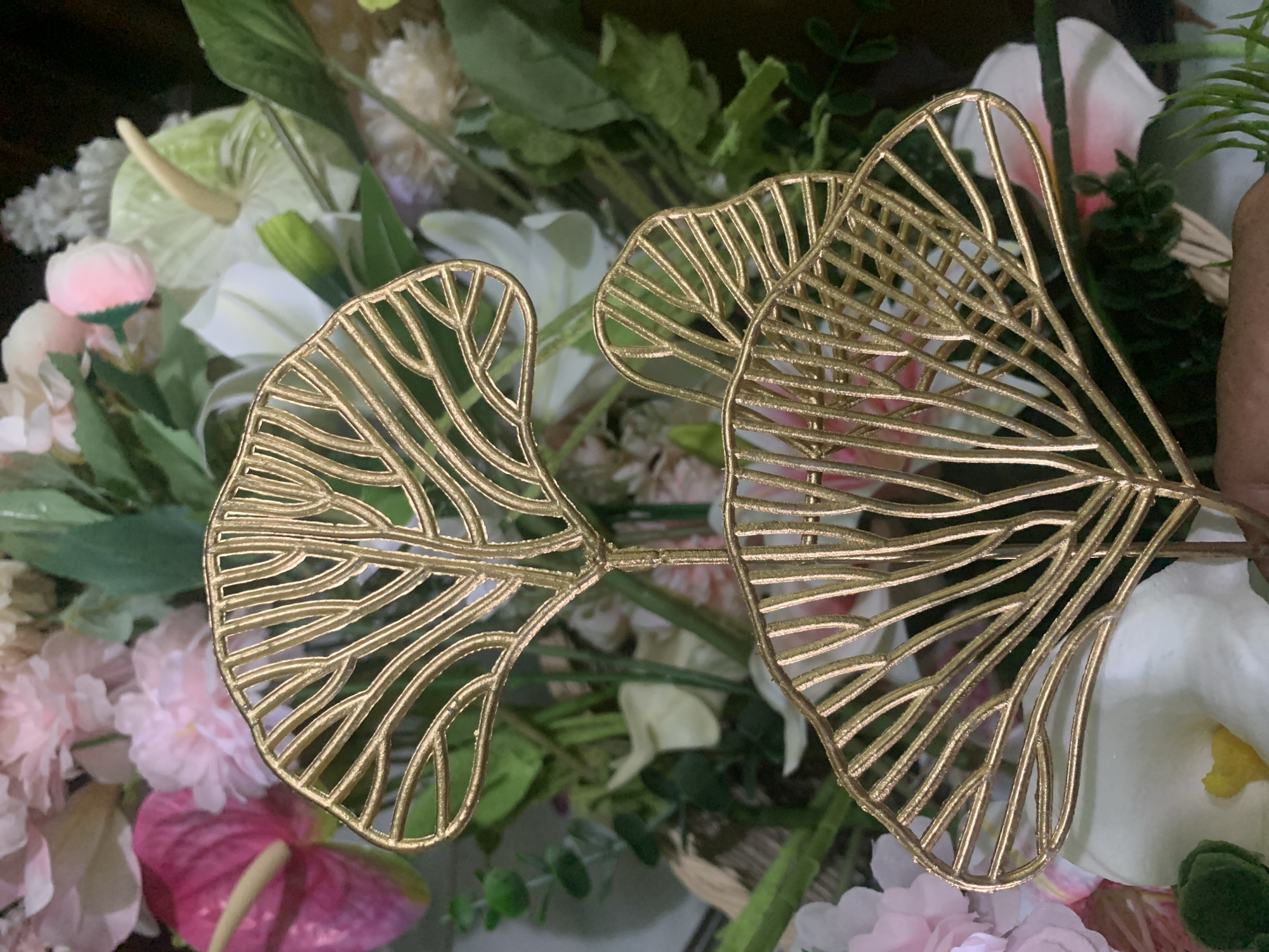 The image displays two golden, leaf-shaped decorative ornaments set against a blurred backdrop of artificial flowers in various shades of pink, white, and green. The ornaments are intricate, with a delicate, veined pattern that resembles a fan. The lighting suggests an indoor setting. There is no discernible location in the city of Kuala Pembuang, Indonesia.