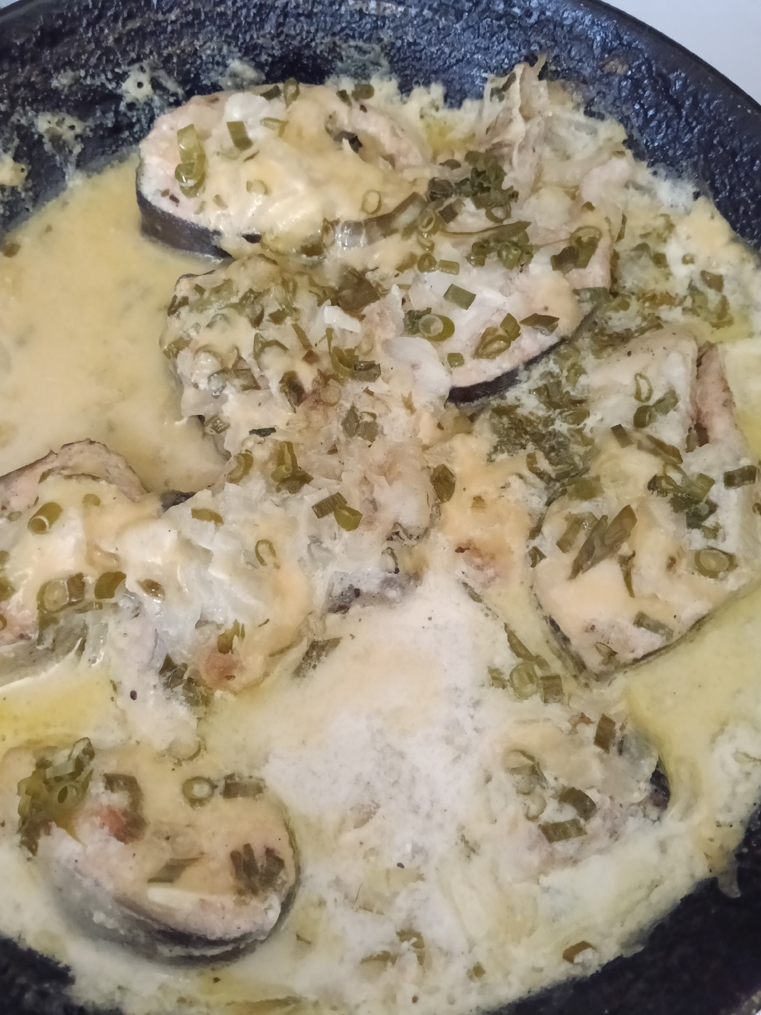 The image shows a pan of cooked fish in a creamy sauce, garnished with green onions. The fish appears to be cut into circular slices. The pan is black and shows signs of use. The image location in the city of Красный Маяк, Russia, is not determinable from the visual information alone.