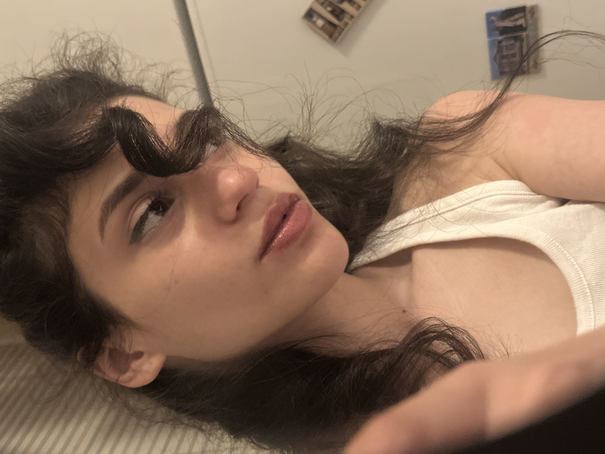 The image shows a close-up, low-angle shot of a young woman with long, dark, wavy hair and fair skin. She is wearing a white tank top. Her head is tilted, and she is looking down and to her left. Her right eye is visible, with dark eyeliner applied. Her lips are slightly parted and appear to have a glossy sheen. A lock of her hair is curled over her forehead. Her left hand is partially visible in the foreground, out of focus. The background consists of a textured wall with two small framed pictures on it, and a surface with horizontal lines, possibly a blind or a wall covering. There is no visible indication of the city of Sofia, Bulgaria, or any other specific location.