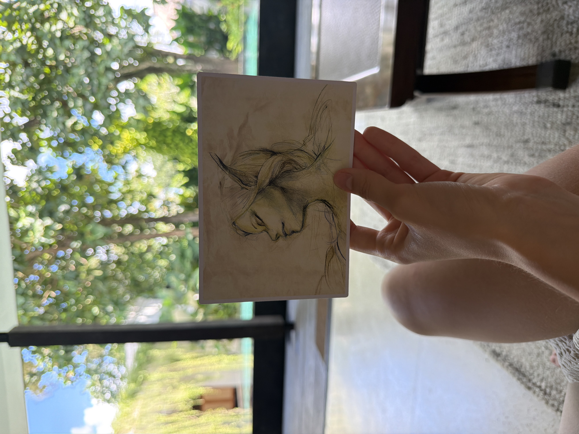 A person is holding a drawing of an elf in their right hand. The drawing is on a small, rectangular piece of paper with slightly rounded corners, depicting a profile of a character with pointed ears and elaborate braided hair. The person's forearm and knee are visible. The background is a bright, out-of-focus scene of trees and sky, as viewed through a window frame. A portion of a chair is visible. Based on information available on the Internet, the location is likely in Mas, Indonesia.