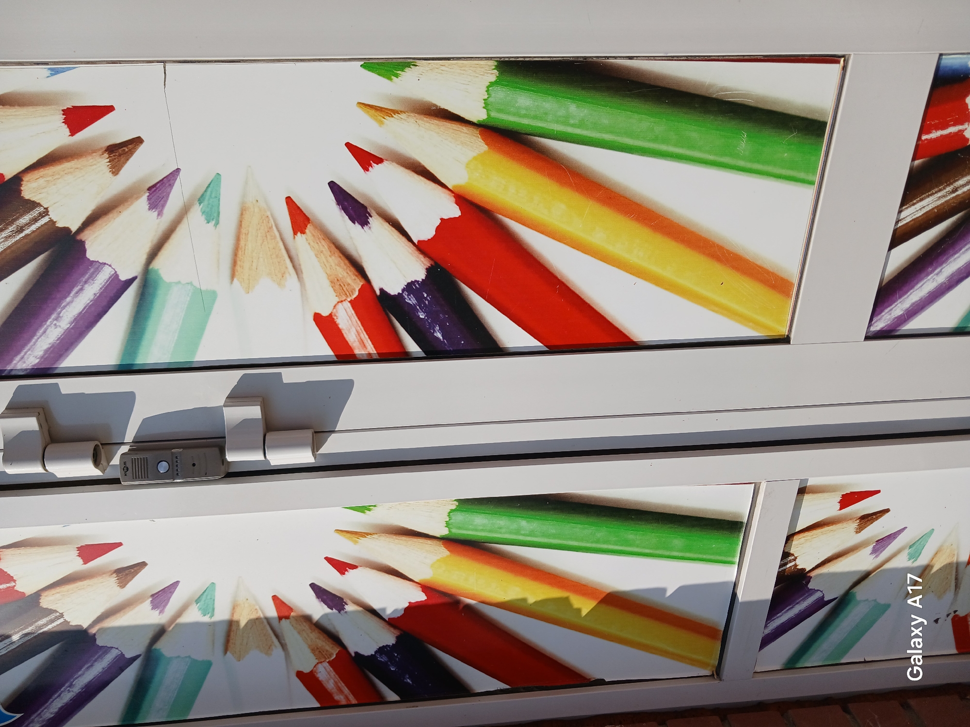The image shows what appears to be a set of display cases or windows featuring images of colored pencils arranged in a semi-circular pattern. The display cases are white-framed, and one of them has a lock mechanism visible. The images of pencils are bright and colorful. The presence of text "Galaxy A17" suggests the image was taken by or with a Samsung Galaxy A17 smartphone. Based on the images, this would be impossible to determine that it is in Yekaterinburg, Russia. It appears to be outside based on the natural lighting. The brickwork is partially visible at the bottom of the frame.