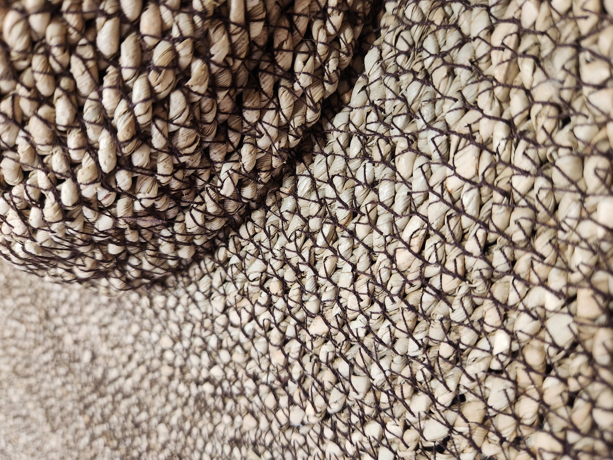 This is a close-up, high-angle shot of a woven textile, likely made from natural fibers such as straw or raffia. The material is woven with a darker, possibly twine-like thread, creating a visible grid pattern. The main subject is the texture and pattern of the weave itself, with two distinct areas of the material visible, one folded over the other, creating a layered effect.
In the upper left portion of the image, the weave is tightly packed and forms a curved surface, suggesting it might be part of a basket, mat, or furniture. The color of the fibers ranges from off-white to light beige, with some darker striations within the individual woven elements. The darker thread used for the weave is a deep brown, providing a strong contrast.
The lower right portion of the image shows a flatter, more spread-out area of the same woven material. The weave appears slightly looser here, and the individual strands are more visible. The lighting creates shadows that highlight the dimensionality of the weave. The background is a blurred continuation of the woven texture, suggesting the object fills the frame.
There are no people, animals, or any discernible activity taking place. The image focuses solely on the material's surface and construction. No text is visible. The setting and time of day are not indicated. The visual emphasis is on the tactile quality and intricate pattern of the woven material.