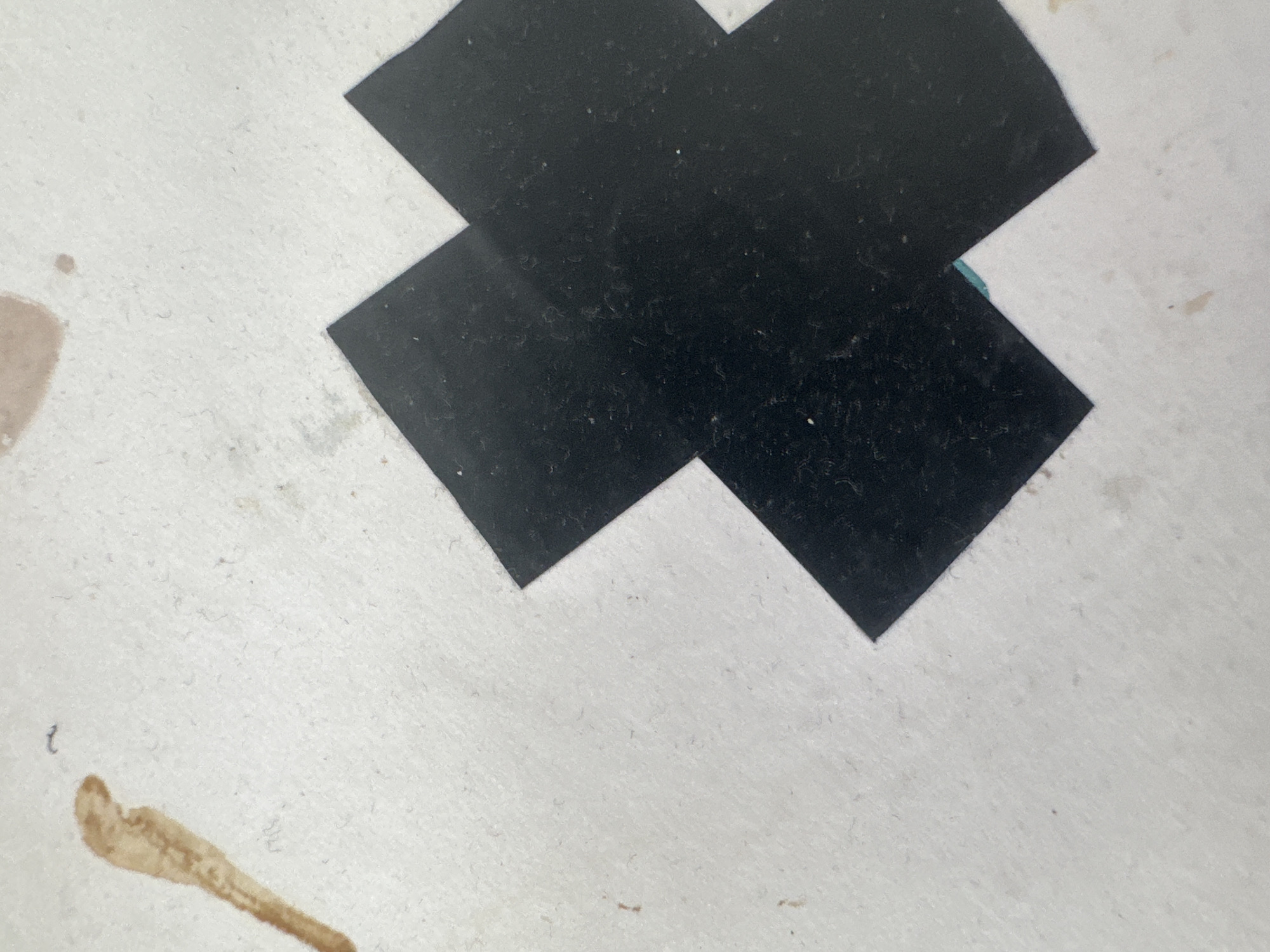 The image is a close-up, top-down view of a white textured surface, possibly paper or a painted wall, marked with a black 'X'. The 'X' is formed by two overlapping rectangular strips of black tape, creating a sharp, angular shape. The tape appears to have a matte finish and is slightly dusty. The white surface itself has some subtle imperfections, including faint brown smudges and a few scattered dust particles. There is a small, faint teal-colored line visible where the two pieces of tape overlap. In the lower left corner, there is a distinct brown, elongated stain or mark. The lighting is even and diffused, suggesting an indoor setting. There is no discernible time of day, weather, or specific location cues beyond the context of Belgrade, Serbia, which is not visually apparent in the image itself. No text is visible in the image. The image focuses solely on the texture of the surface and the stark black marking.