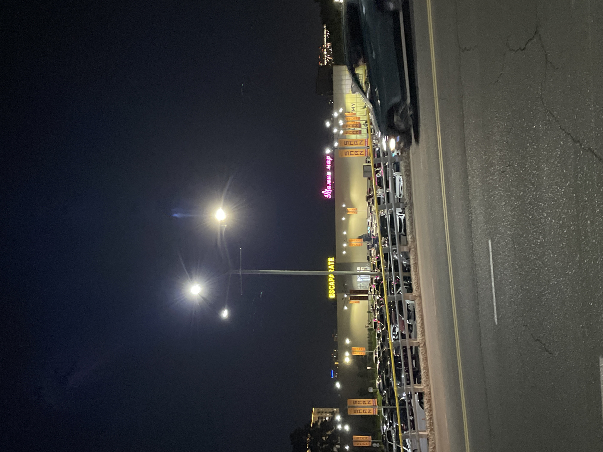 The image captures a nighttime scene in Almaty, Kazakhstan, likely in the vicinity of a shopping center. In the foreground, a dark asphalt road with yellow dividing lines is visible, with a slight crack in the surface. Beyond the road, a parking lot filled with numerous cars is illuminated by overhead lights and the signage of a building.
The building's facade features prominent illuminated signs, including "ESCAPARATE" in yellow capital letters and another sign with Cyrillic script that appears to read "МАНУЛ МИР". Several vertical banners with Cyrillic text, possibly brand names, are also displayed. The overall lighting suggests a commercial area.
In the mid-ground, a dark-colored SUV is driving past the parking lot, its headlights and taillights illuminated. The sky above is dark, with streetlights casting a bright glow. The scene conveys a bustling commercial atmosphere, even at night. The presence of multiple cars and illuminated signs suggests ongoing activity within the shopping center and its surroundings.