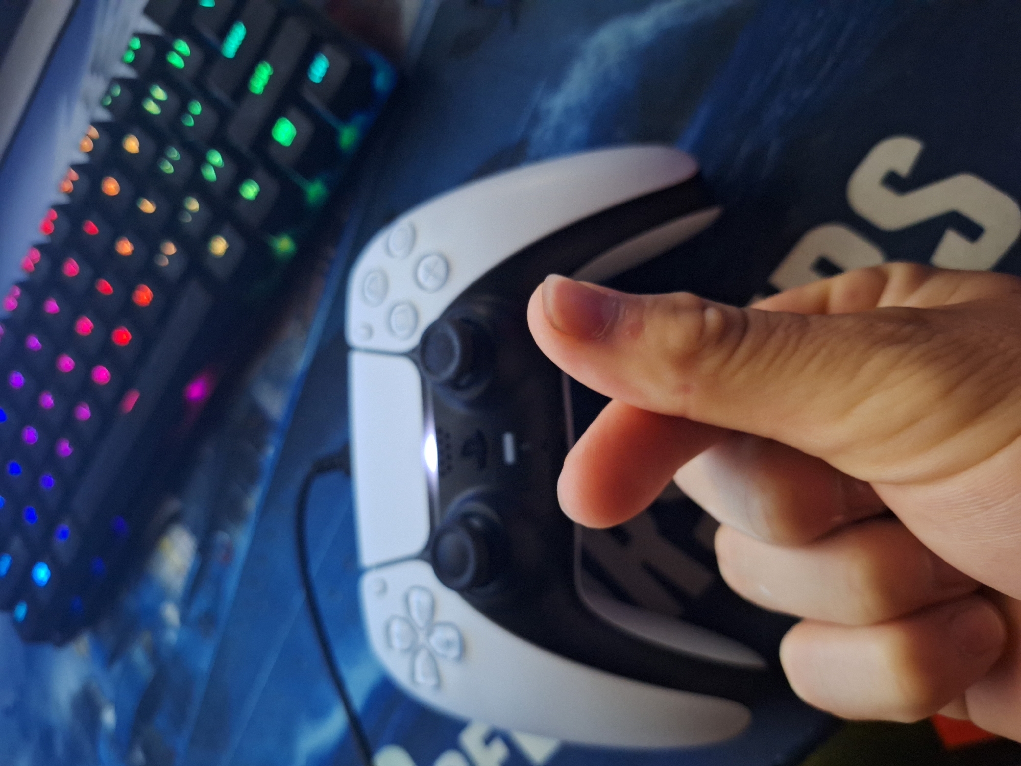 In Varna, Bulgaria, a person's hand, positioned in the foreground, forms a Korean heart gesture with their thumb and index finger. Behind the hand, a white and black PlayStation 5 controller is visible. The controller is resting on a blue mat that has some white text printed on it. In the background, a dark mechanical keyboard with colorful RGB lighting is partially in view. The overall scene suggests a gaming setup.