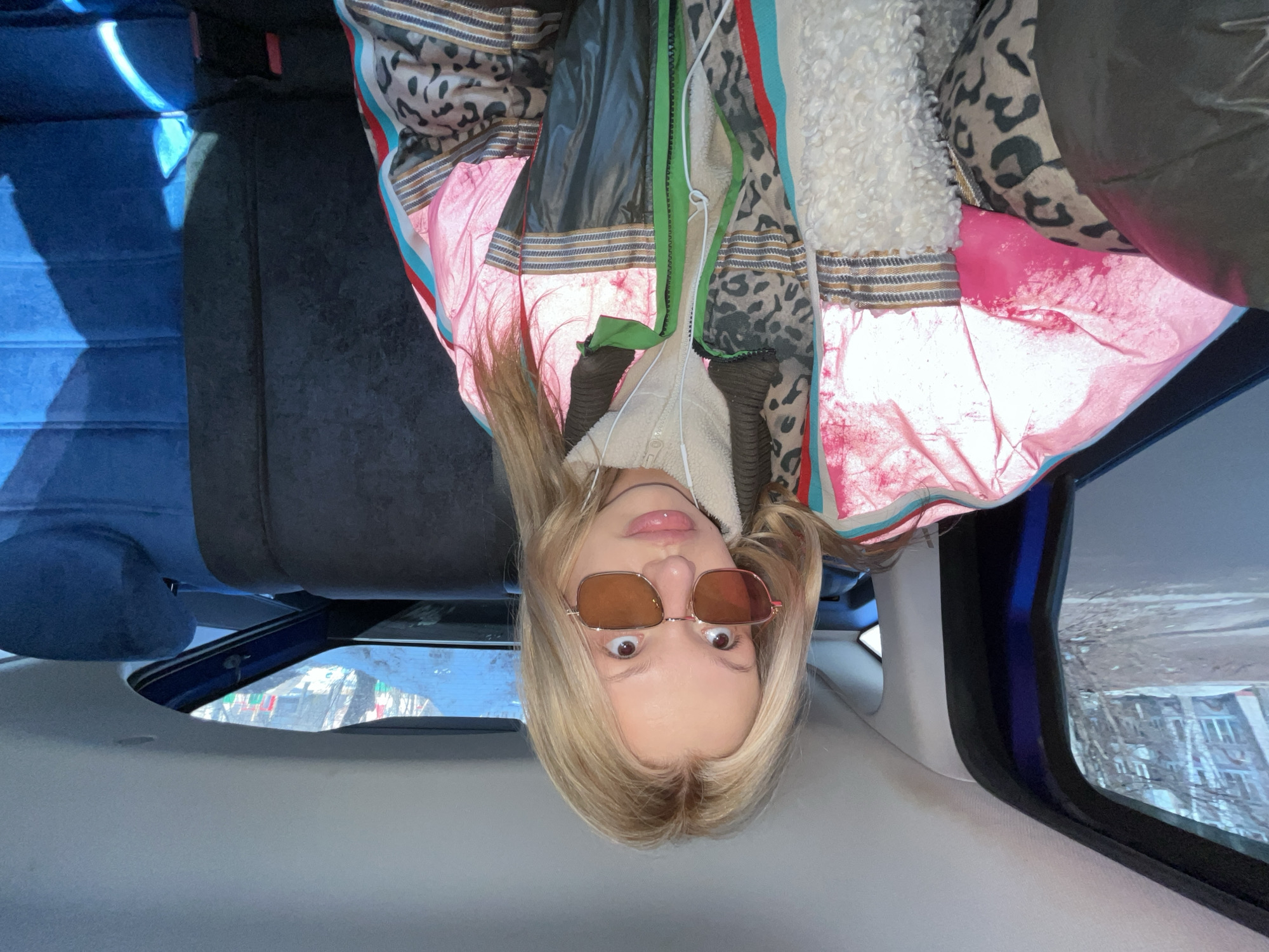 A young adult female is seated in the rear passenger compartment of a vehicle, angled slightly towards the camera as if taking a selfie. She has long blonde hair and fair skin, with a labret piercing on her lower lip. She wears amber-tinted rectangular sunglasses with thin rose-gold frames, looking upwards and slightly to her right. White wired earphones are visible around her neck and chest. Her attire consists of multiple layers: an inner cream-colored fleece jacket and an outer multi-panel jacket featuring reflective pink sections, leopard print patterns, dark grey glossy material, and striped bands in blue, red, white, gold, and black. A green zipper element is visible on the outer jacket.
The car's interior shows a grey headliner and dark grey fabric seats. A blue fabric seat and headrest are visible in the front passenger area. Through the left rear window, an exterior scene unfolds: a paved road or street with patches of snow or bare ground, deciduous trees without leaves, and a light-colored multi-story residential building with visible windows. Through the rear window, more bare trees and snow-covered ground are visible, along with distant, partially obscured colorful elements resembling flags or banners (red and green).
The exterior environment, visible through the vehicle's windows, displays a typical urban residential area in a cold climate, consistent with Chaikovsky, Russia. Specific landmarks to pinpoint an exact location within the city are not discernible from the image.