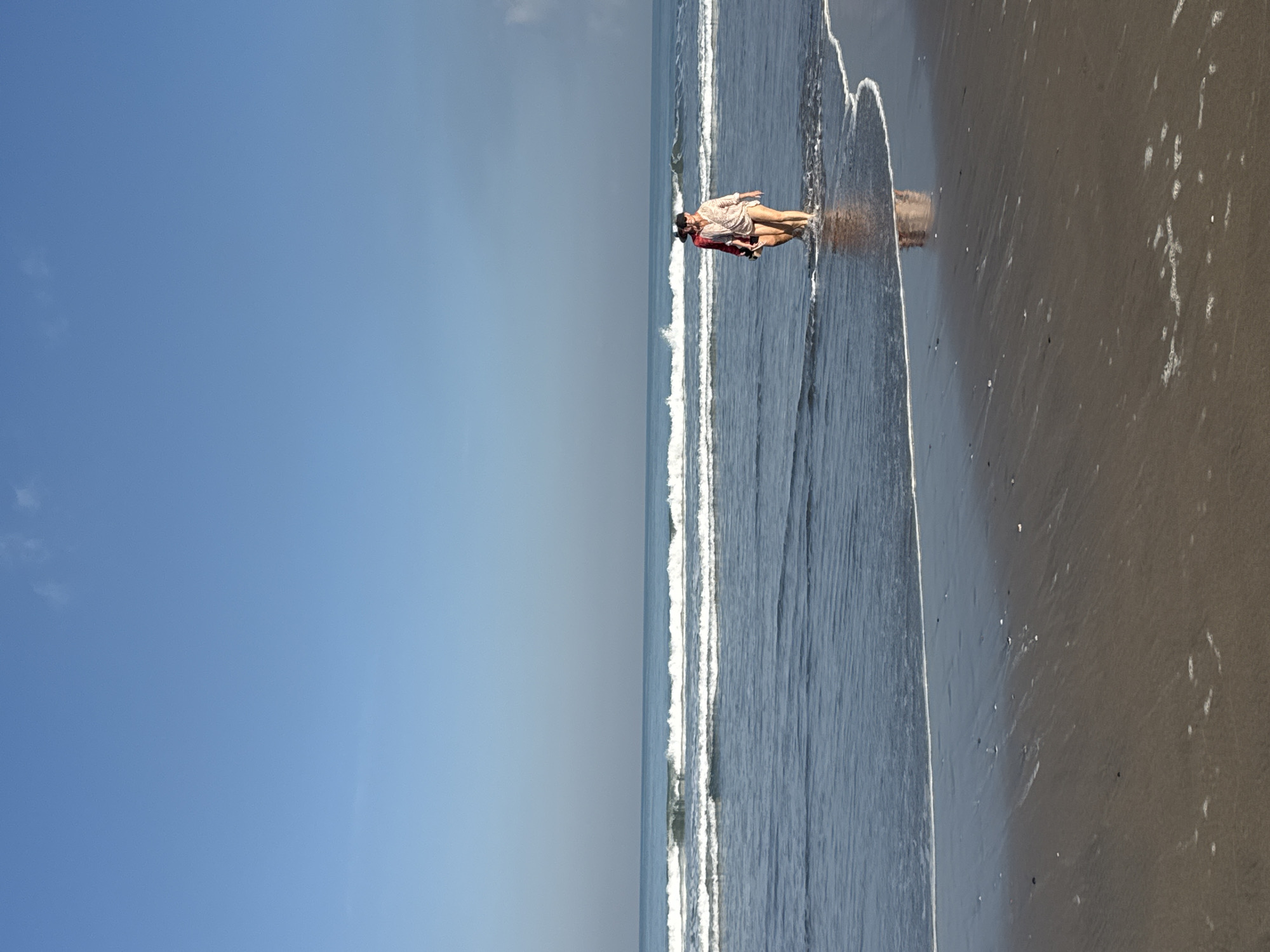 The image captures a serene beach scene in what appears to be Kuta, Indonesia, during daytime under a clear, bright blue sky with only a few wispy clouds. The composition is divided horizontally into the expansive sky, the ocean, and the wet sandy shore.
The central subject is a single person, likely a woman, standing near the water's edge. She is wearing light-colored, patterned clothing, possibly a flowy top or dress over shorts, and a dark hat. Her reflection is distinctly visible in the shallow, still water and wet sand, suggesting calm conditions. She appears to be standing still, possibly enjoying the view or the sensation of the water.
Gentle white waves are breaking and receding along the shoreline, creating white foam lines that extend across the frame. The ocean water is a deep blue, transitioning to lighter hues near the horizon. The sand on the beach is dark and wet where the waves touch it, showing some texture and small ripples from the water movement. Further up, the sand appears slightly drier and less reflective.
The overall atmosphere suggests a peaceful and pleasant beach outing. There is no visible text in the image.
