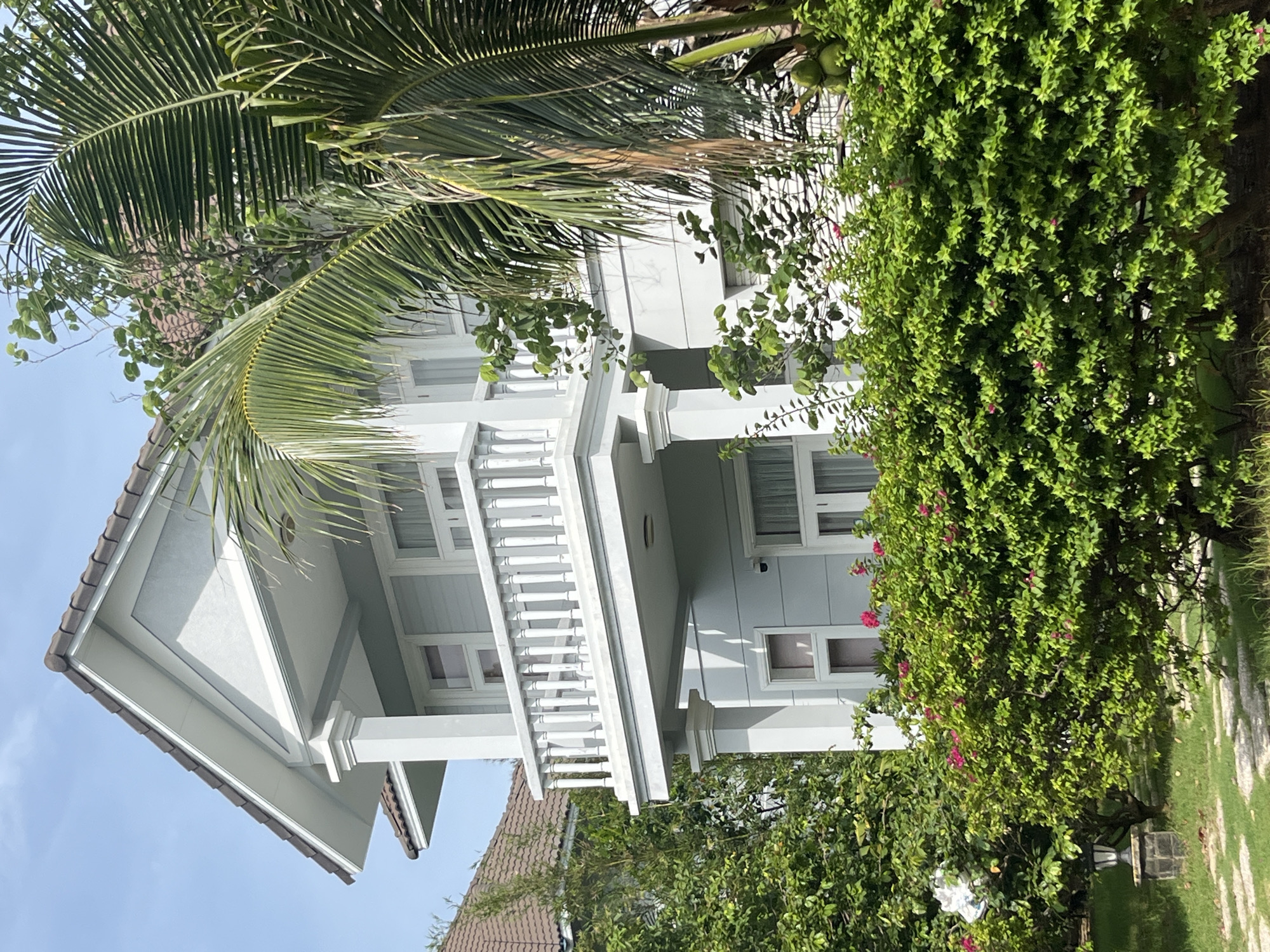 The image shows a two-story light blue house with white trim and a white balcony railing on the second floor. The house has multiple windows and a gabled roof with brown tiles. The house is surrounded by lush green foliage, including a large palm tree on the right side of the frame and flowering bushes in the foreground. A stone pathway with stepping stones is visible in the lower left corner, next to a lantern. The scene appears to be a residential property, likely in a tropical or subtropical climate, given the presence of the palm tree and dense vegetation. The sky is clear and blue, suggesting a sunny day. The image's composition and focus on the house and its surroundings suggest it could be part of a real estate listing or a travel brochure for a location like Phu Thuy Ward, Vietnam, known for its tropical climate and scenic beauty.