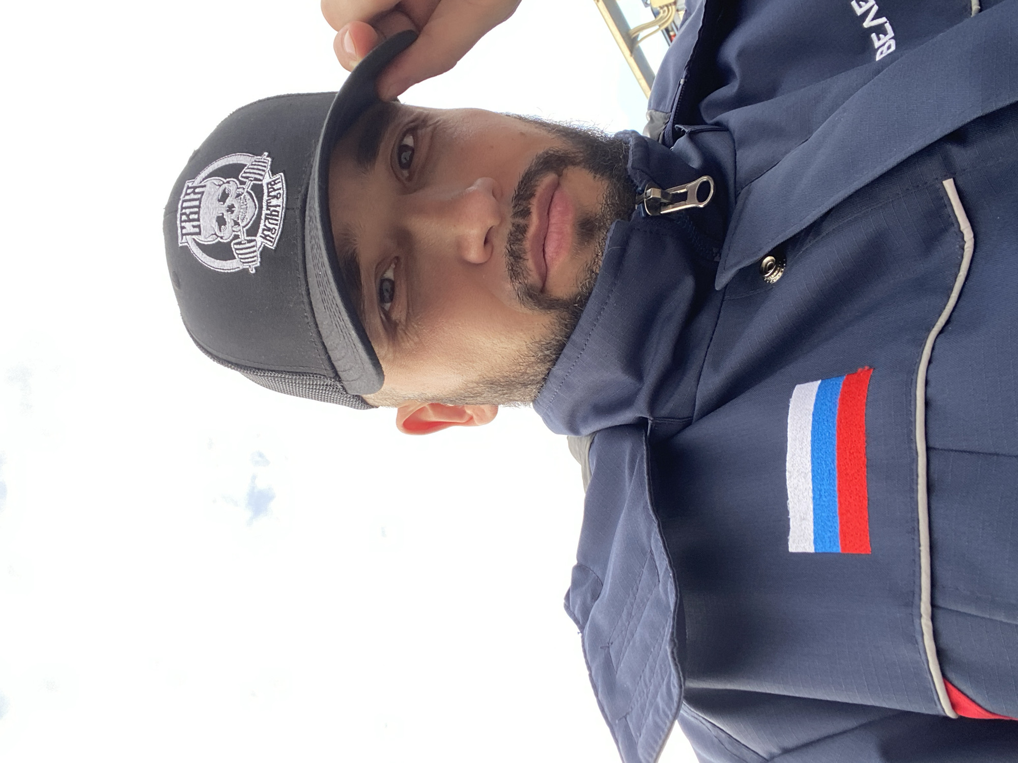 A close-up, angled portrait features a man looking directly at the camera. He wears a dark blue jacket and a black baseball cap. The man has a beard and mustache, and his expression is neutral. His right hand is visible, holding the brim of his cap.
The black baseball cap has a white embroidered logo on the front: a stylized skull holding a barbell, encircled by text. The text above the skull reads "СИЛА" (SILA, meaning "Strength" or "Power"), and the text to the right of the skull reads "КУЛЬТУРА" (KULTURA, meaning "Culture"). The cap also has a grey mesh back.
The man's dark blue jacket has a silver zipper pull and white trim details. On the upper left chest of the jacket, there is a rectangular patch depicting the flag of Russia, with horizontal stripes of white, blue, and red. Partially visible white embroidered text on the jacket collar reads "BEA".
The background is a bright, overexposed white, suggesting an outdoor setting under a clear, bright sky, likely during the daytime. The location context provided is Koskolovo, Russia.