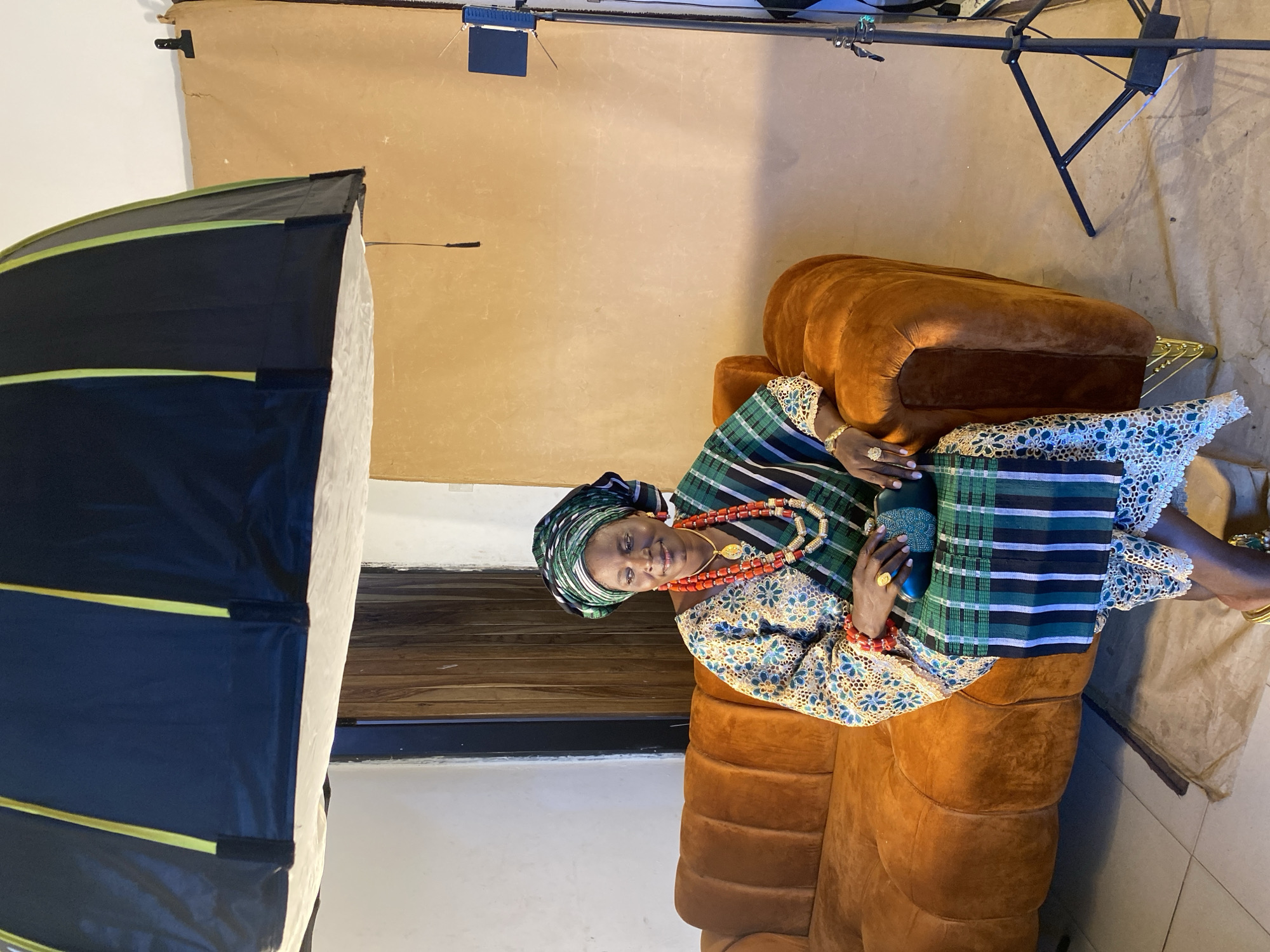 A middle-aged woman in traditional Nigerian clothing sits on an orange couch in what appears to be a photography studio. She is wearing a green and black patterned headwrap, a floral-patterned blouse, and a striped green and black sash. She is also wearing coral beads around her neck and wrists, and gold jewelry on her fingers and wrists. She is holding a small blue purse in her hands. Behind her is a tan backdrop. Lighting equipment, including a softbox, is visible in the background. There is no evidence in the image to confirm it is taken in Bukuru, Nigeria.