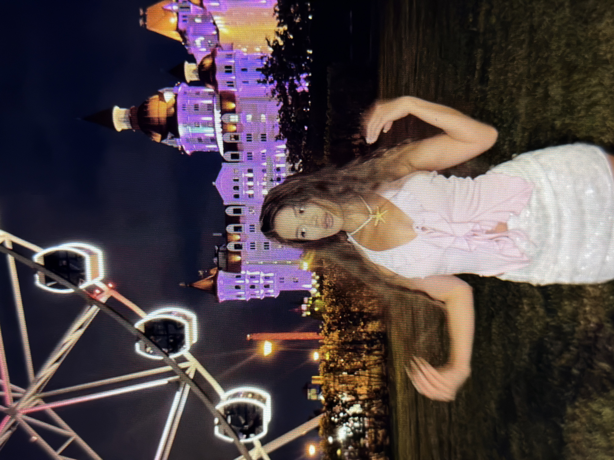 The image depicts a young woman standing in a grassy area at night, with a Ferris wheel and a brightly lit, ornate building in the background. The woman is wearing a pink ruffled top and a white sparkly skirt. Her arms are raised, and her body is angled slightly to the side. Her long, dark hair is down and appears to be blowing in the wind. The building behind her is illuminated with purple and orange lights, resembling a castle or a grand hotel. The Ferris wheel, also lit up, is to the left of the frame. The ground is covered in grass, and there are some trees and bushes visible. The overall atmosphere is festive and perhaps part of an amusement park or entertainment district. The location is the city of Sirius, Russia, known for its modern architecture and entertainment facilities, including those associated with the Olympic Games.