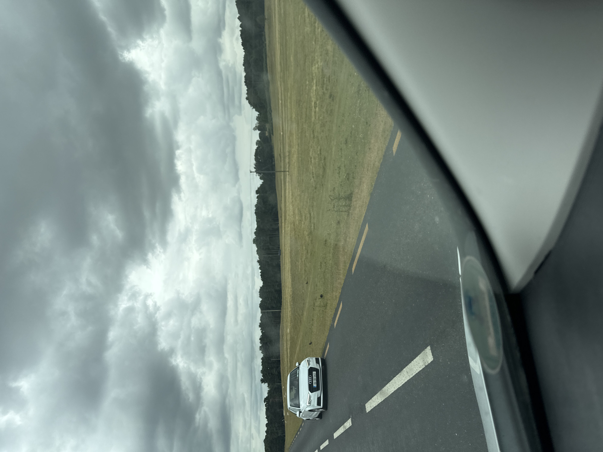 A white Audi is driving on a road in Lumville, Ireland. The sky is overcast with dark clouds, suggesting it may rain. The road is asphalt and has a yellow line on the right side and a white line on the left. The car has its headlights on. The surrounding area is a grassy field with some trees in the distance. The scene appears to be a rural or suburban area outside of Lumville, Ireland.